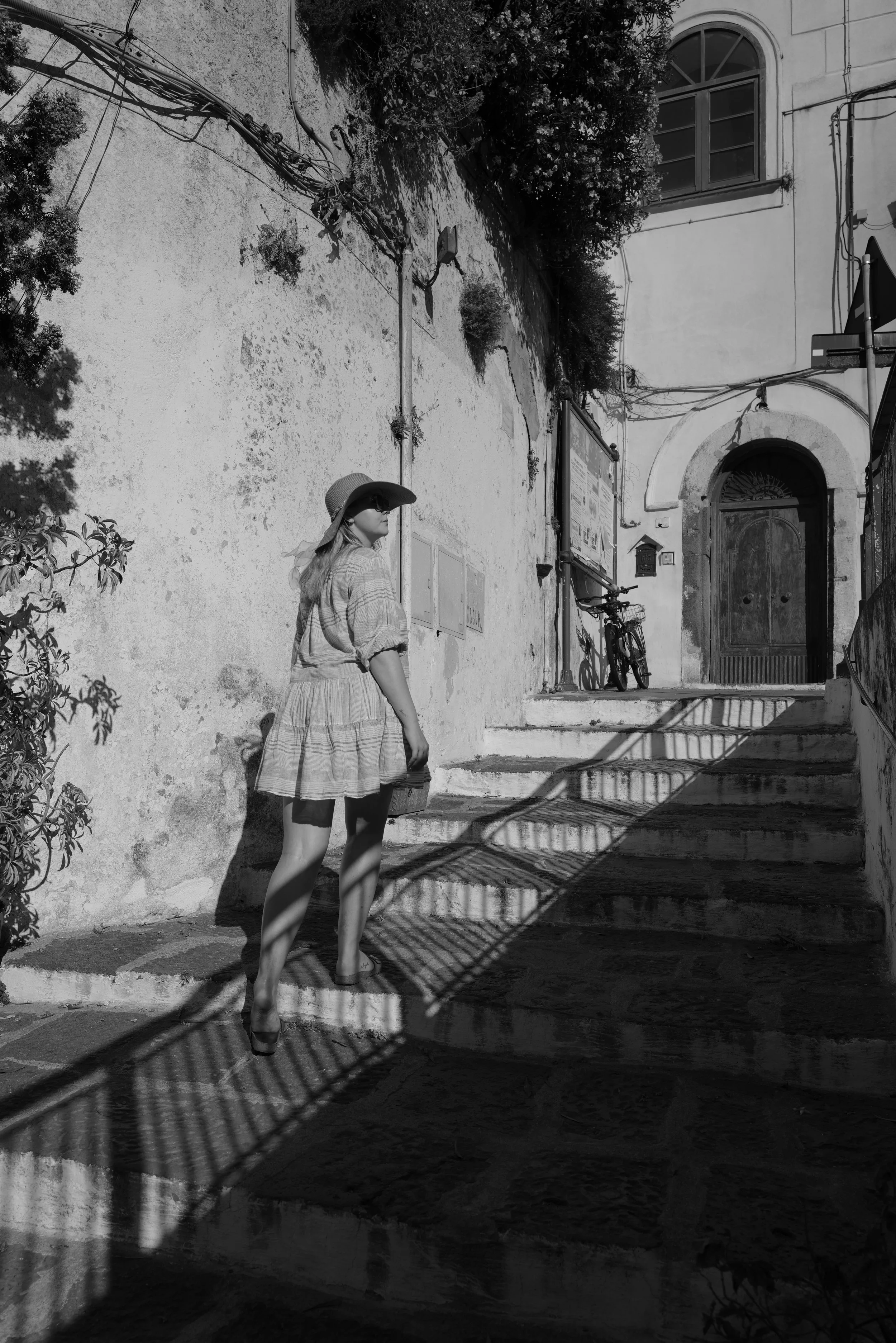 Frau in Sommerkleidung und Hut geht eine Steintreppe hinauf, bei Sonnenlicht; alte Gebäude und Fahrrad im Hintergrund.