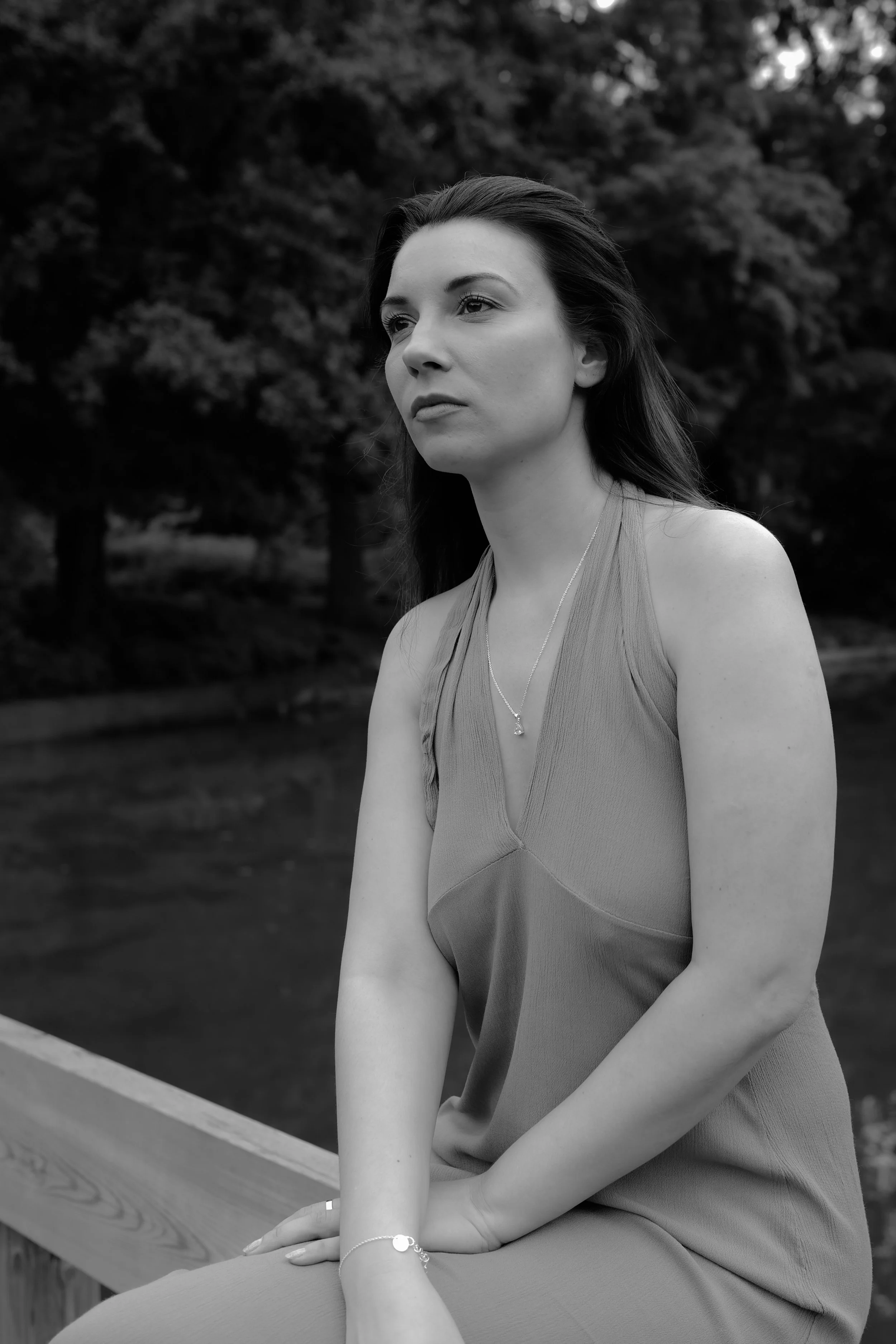 Schwarz-weiß-Foto einer jungen Frau mit dunklen, langen Haaren, die auf einer Holzbrücke sitzt, mit Bäumen im Hintergrund.