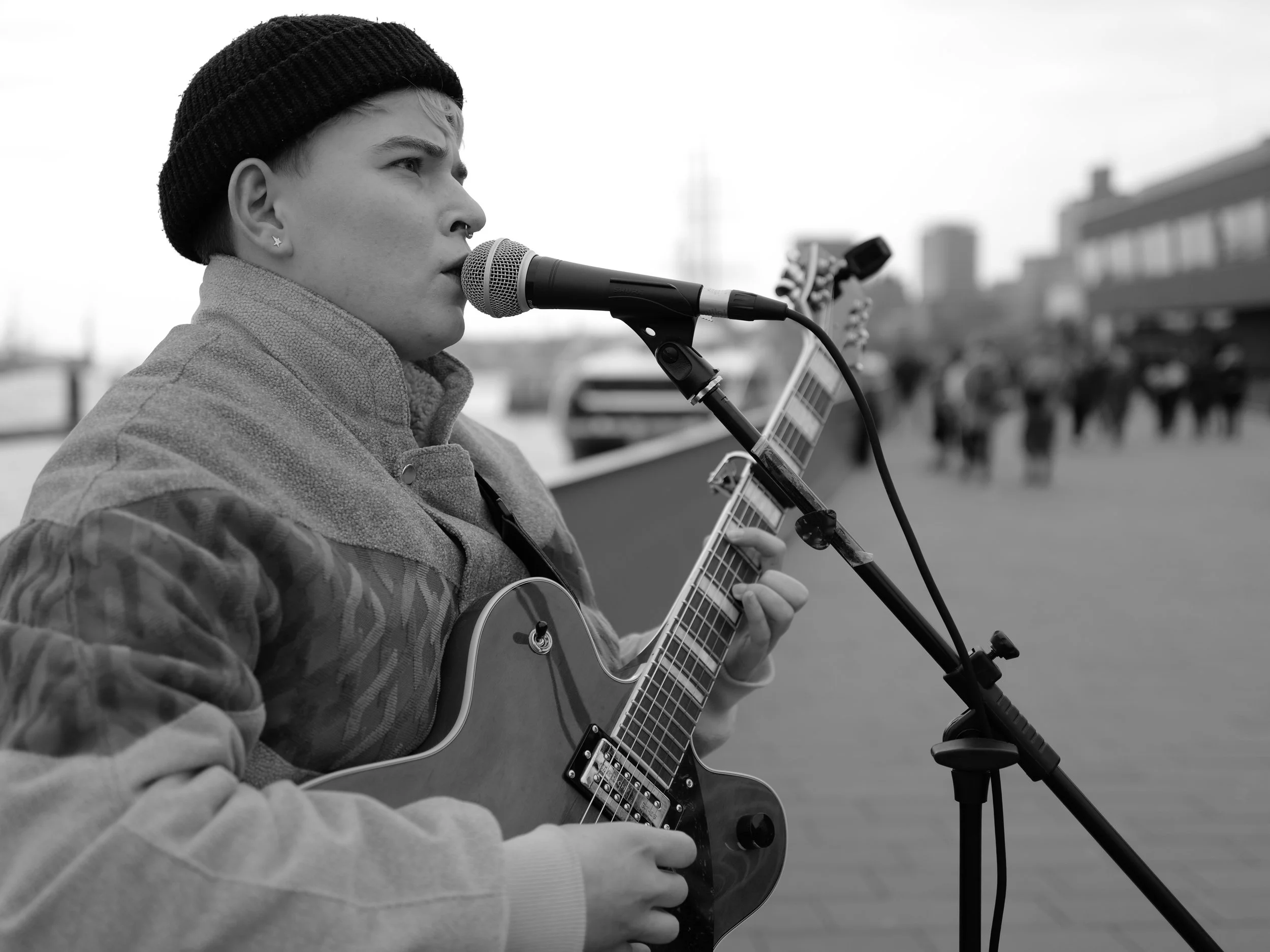 Straßenmusiker mit Gitarre und Mikrofon, schwarz-weiß, spielt Musik im Freien. Im Hintergrund unscharf sind Personen und Gebäude sichtbar.