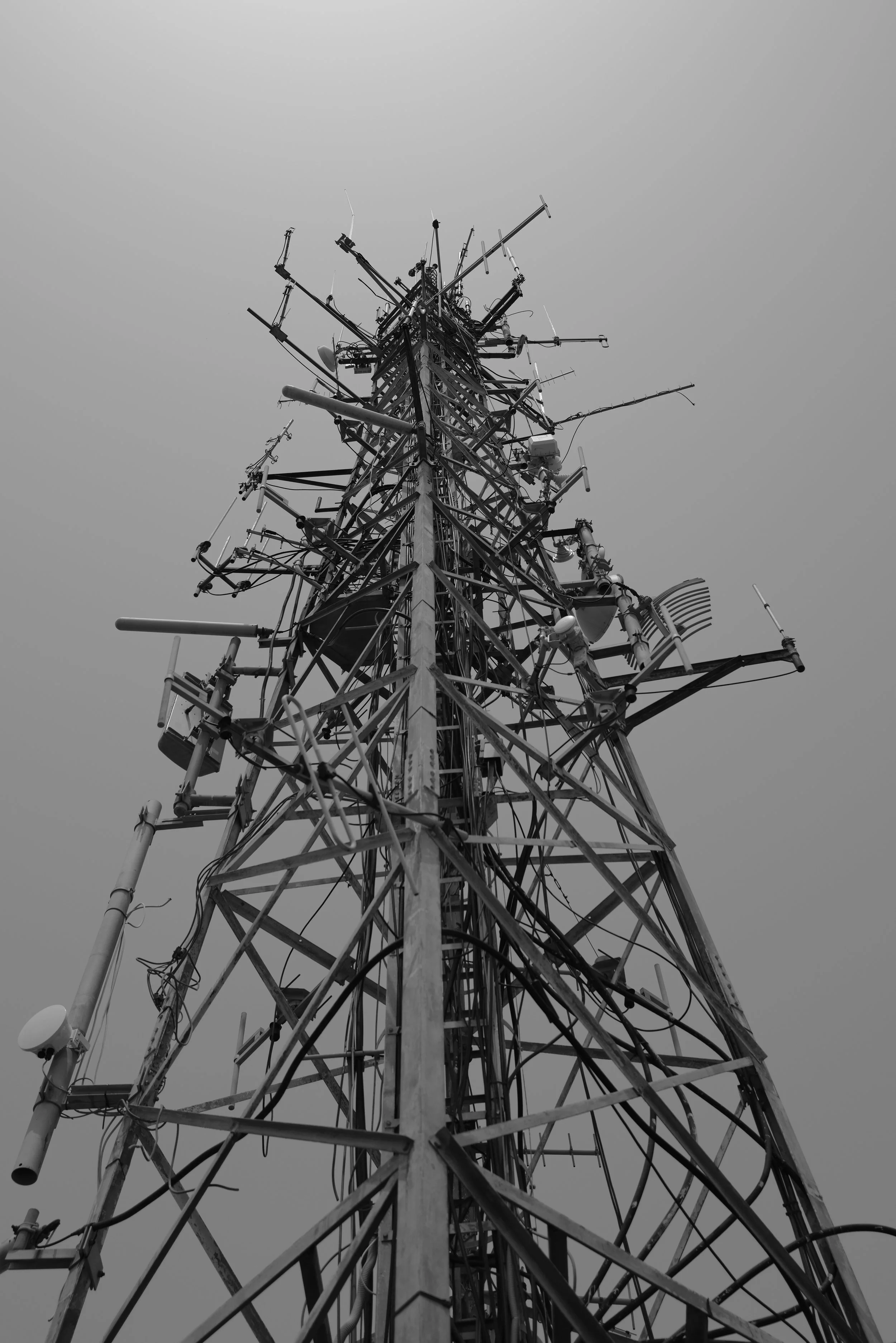 Schwarzweiß-Aufnahme eines hohen Sendemasts mit verschiedenen Antennen und Kabeln gegen den Himmel.