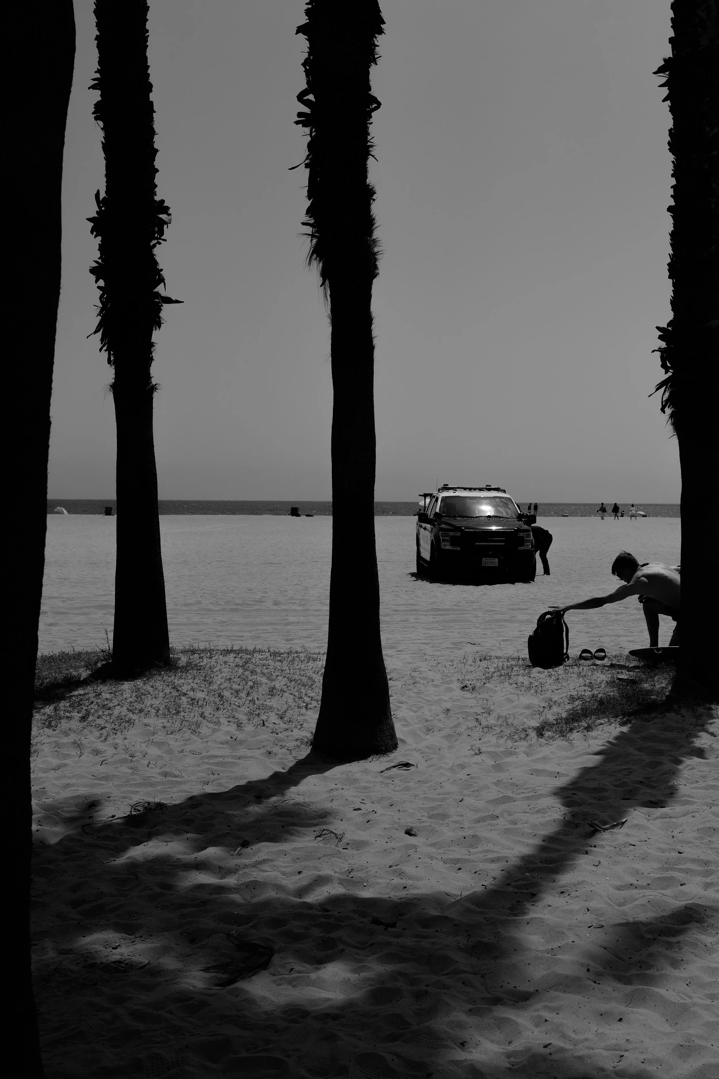 Dünen an einem Strand mit drei Palmen im Vordergrund, einem Wasserrettungswagen, einem Menschen, der sich im Sand bückt, und wenigen weiteren Personen in der Ferne am Wasser.