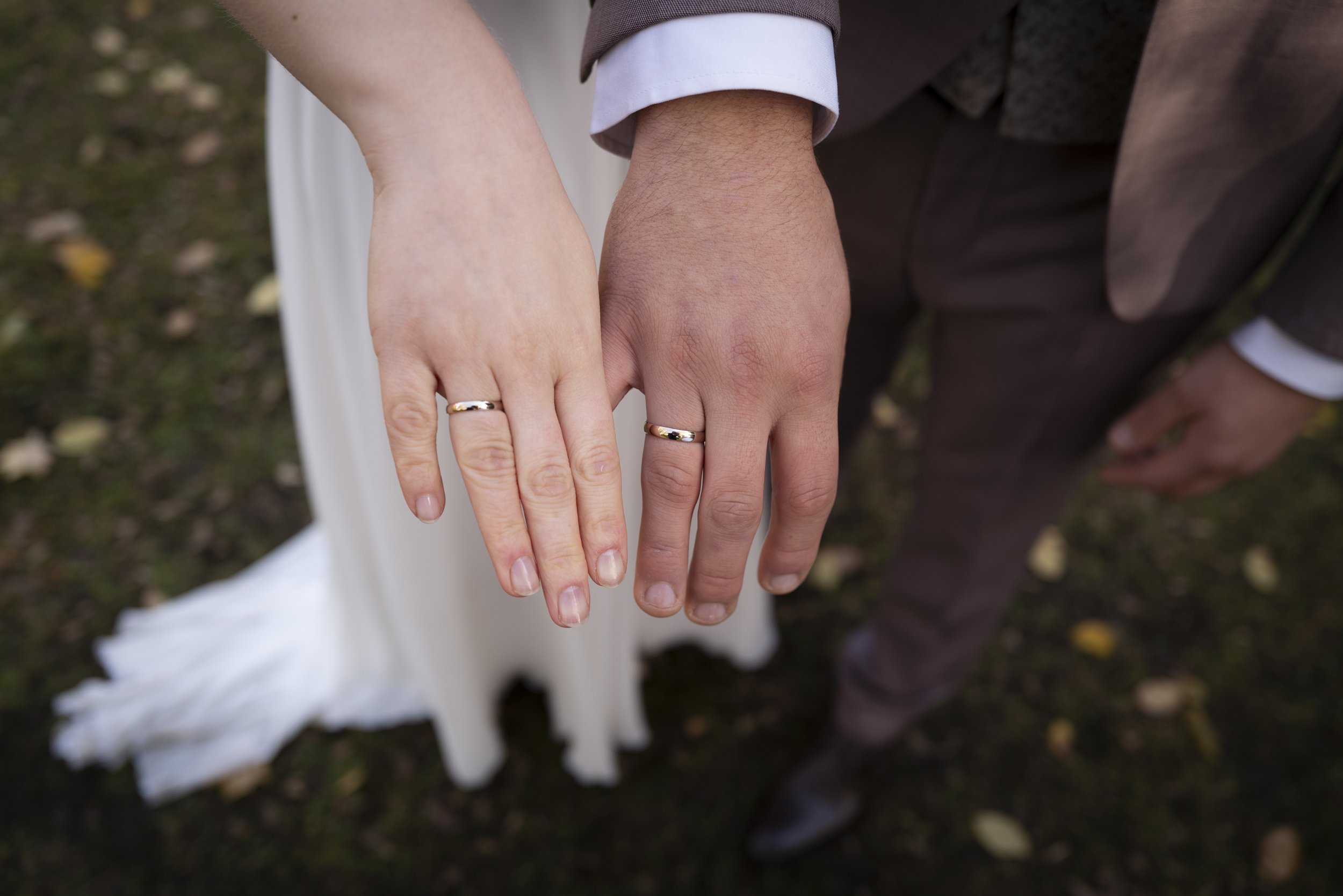 Ein Brautpaar zeigt ihre Ringe, die Hand des Mannes ist näher am Bildschirm, die Hand der Frau ist oben. Das Paar hält sich an den Händen, auf dem Boden liegen Herbstblätter.