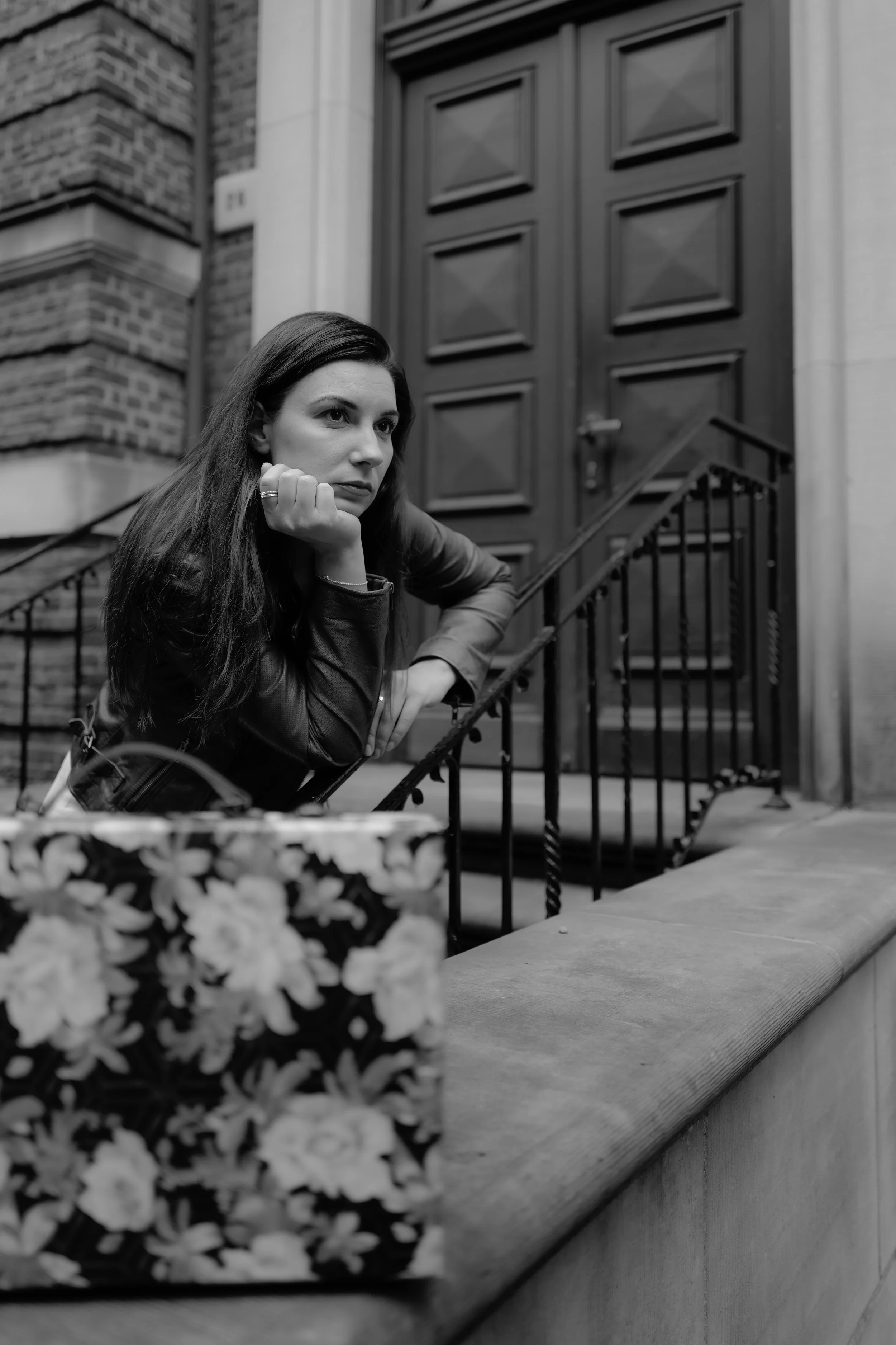 Schwarz-weiß-Foto einer Frau, die auf einer Treppe vor einem großen Holzportal steht, mit nachdenklicher Haltung, trägt eine Lederjacke.
