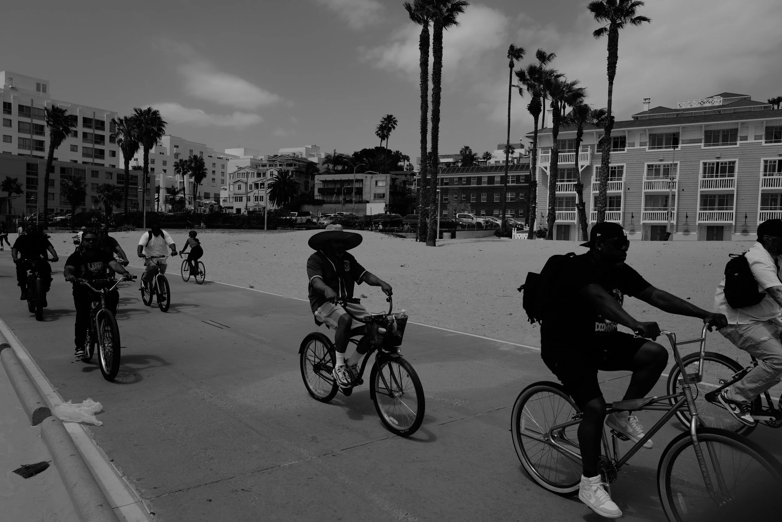 Mehrere Personen fahren Fahrrad an einem sonnigen Tag entlang eines Pfades mit Sandstrand und Gebäuden im Hintergrund, darunter Palmen und Apartmenthäuser.