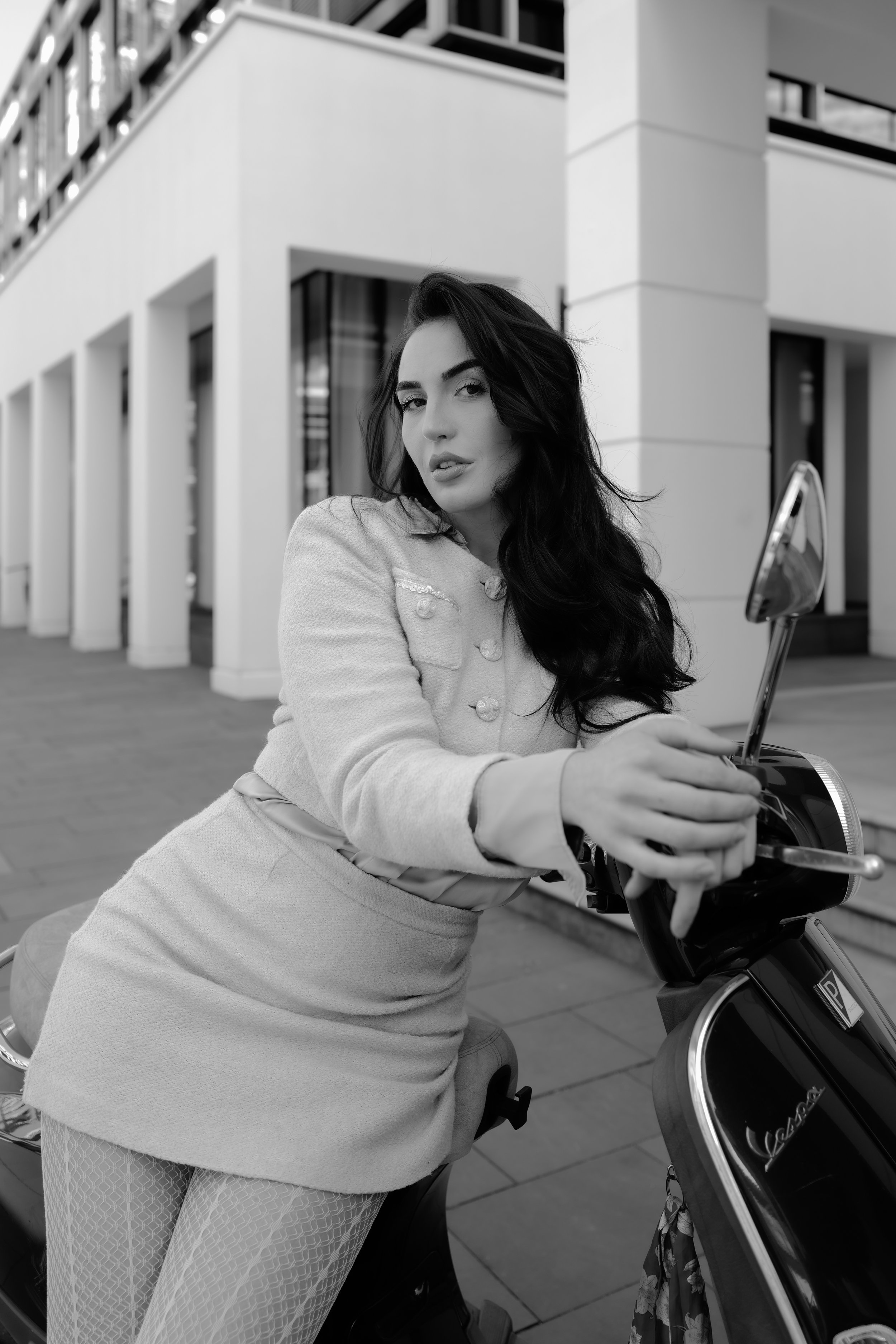 Eine Frau mit dunklen, lockigen Haaren und einem hellen Kleid sitzt auf einem mopedartige Fahrzeug und posiert vor einem modernen Gebäude.