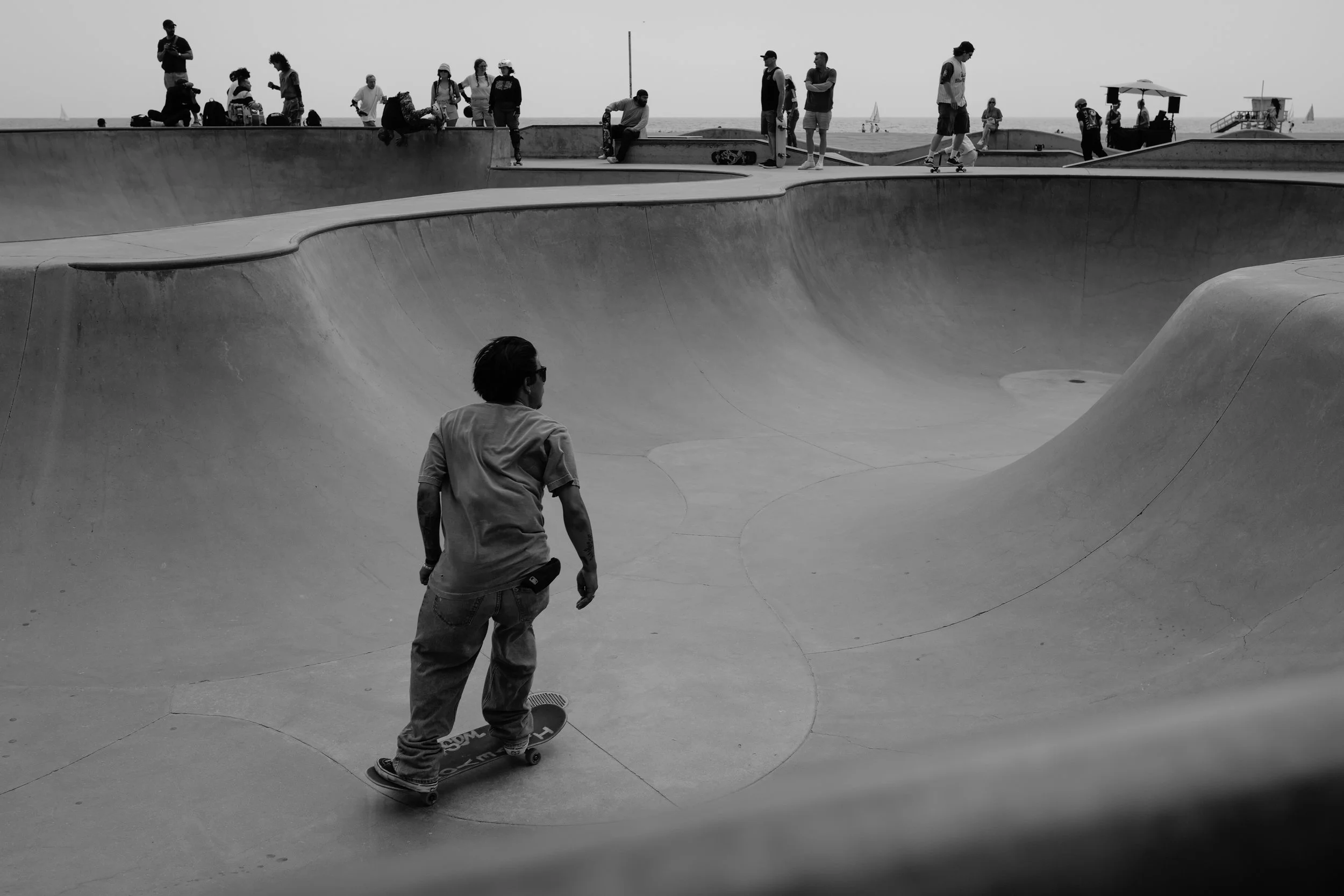 Ein Skateboarder beim Skaten in einem Betonkiefern im Park am Strand, mit mehreren Menschen im Hintergrund, die auf einer Mauer sitzen oder stehen.