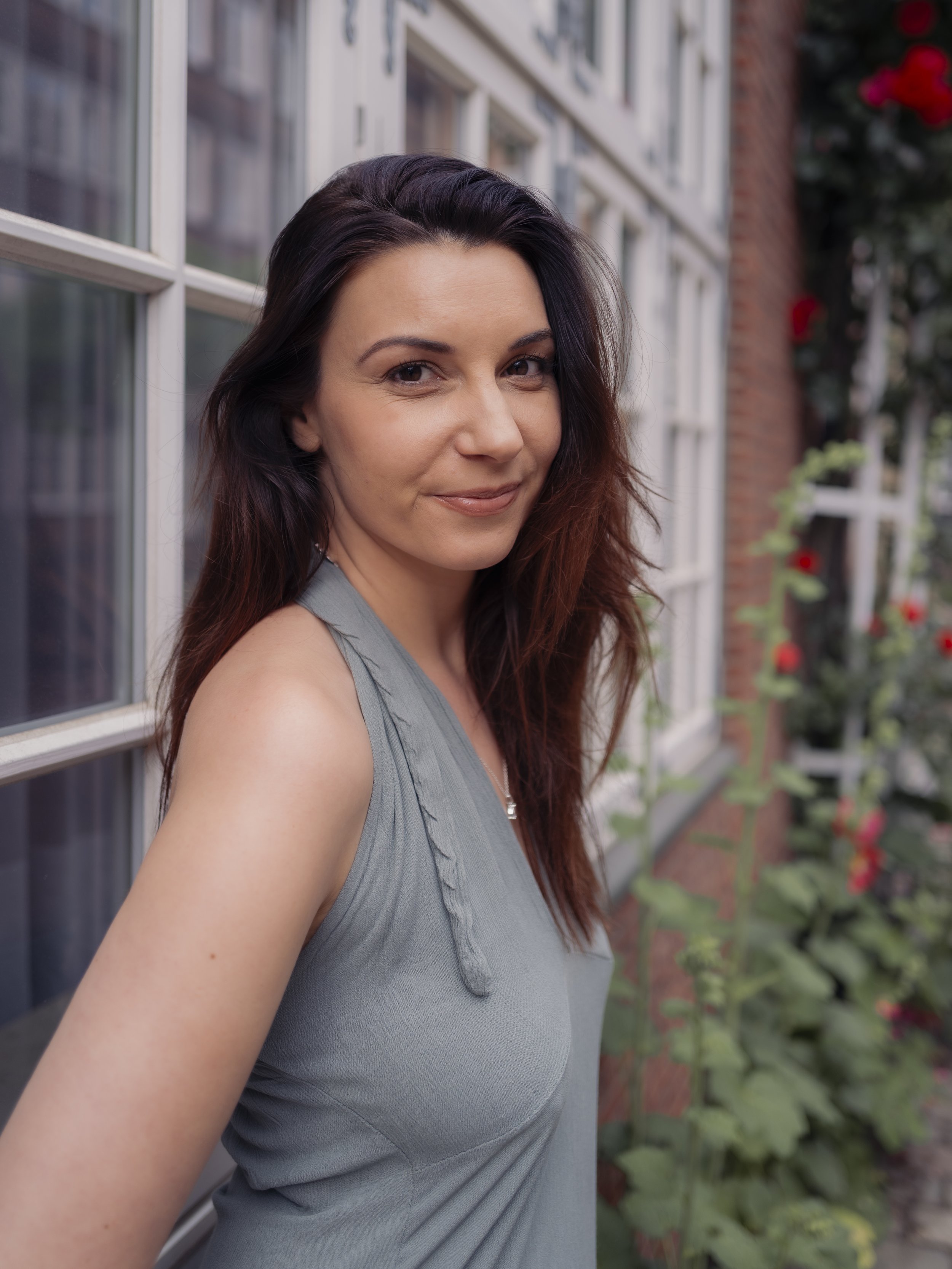 Eine junge Frau mit braunen, gewellten Haaren, trägt ein graues Kleid und steht neben einem Fenster mit weißen Rahmen. Im Hintergrund sind blühende Pflanzen und eine rote Backsteinmauer sichtbar.