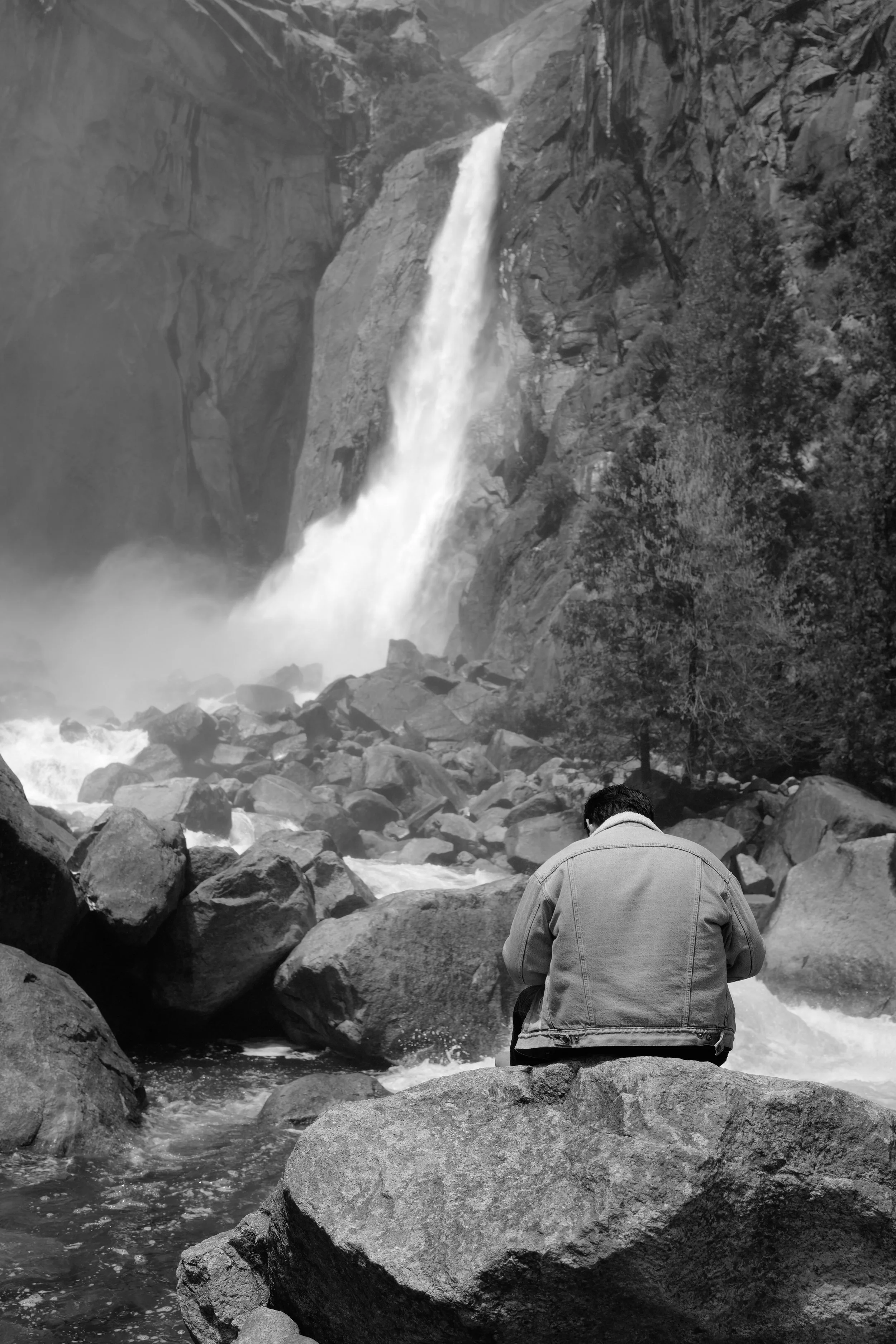 Schwarz-weiß-Foto eines Mannes, der auf einem Felsen am Fluss sitzt, mit einem Wasserfall im Hintergrund.