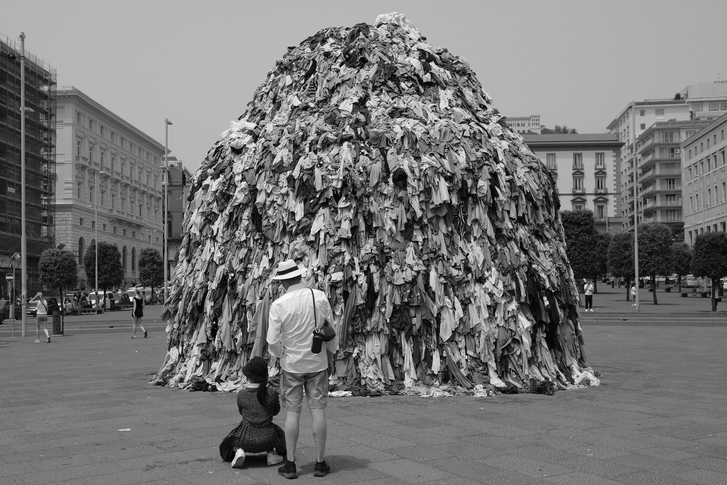Ein großer Haufen aus Kleidung in der Mitte eines Platzes, umgeben von Gebäuden. Zwei Personen betrachten die Installation.