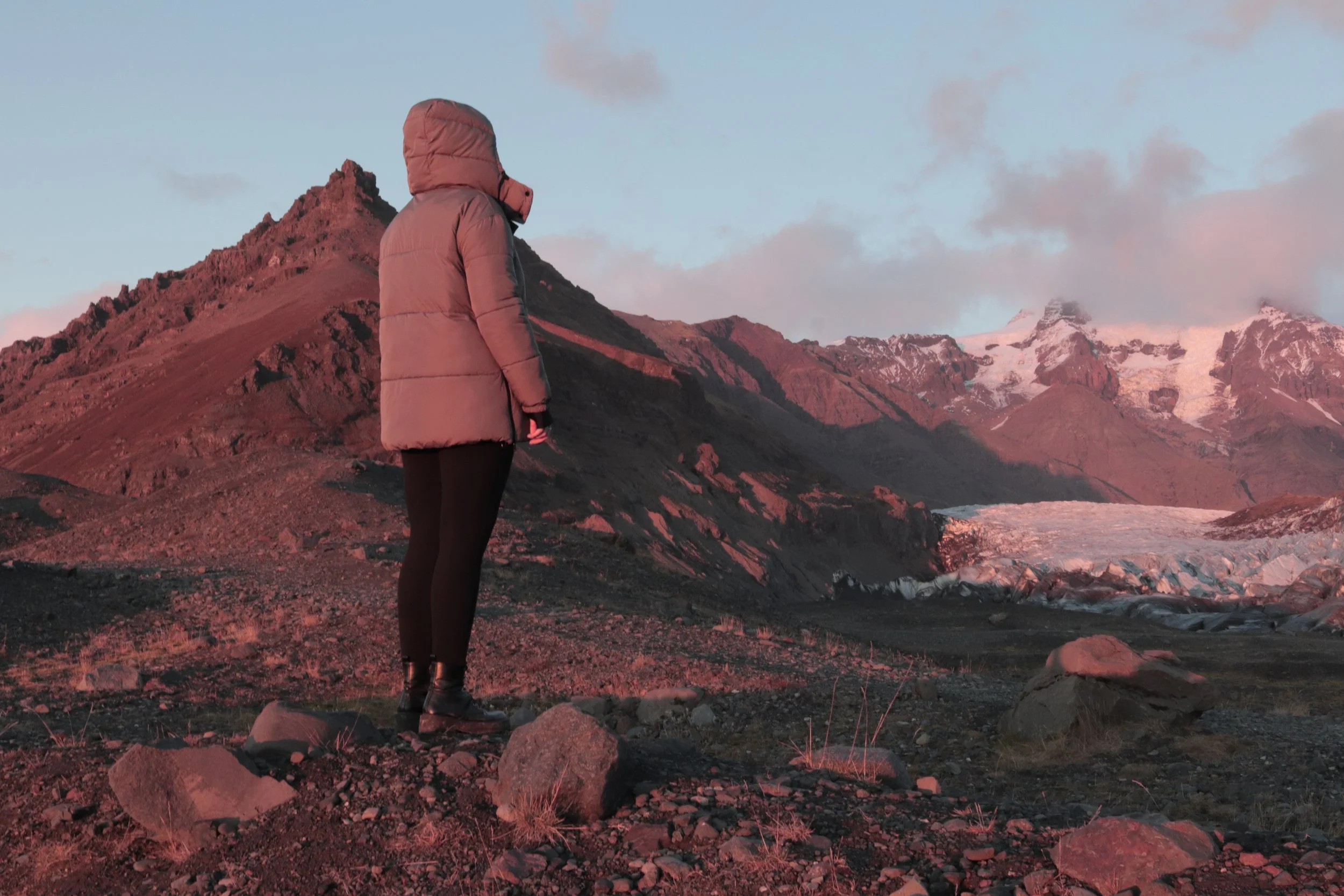 Person in warmer Kleidung steht vor einer vulkanischen Berglandschaft mit einem Gletscher im Hintergrund bei Sonnenuntergang.