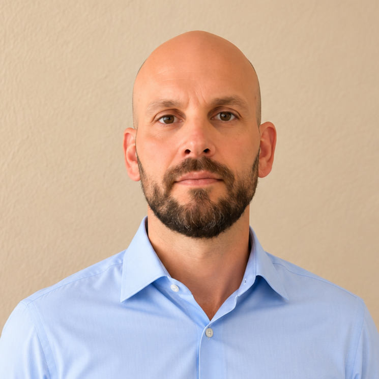 Portrait of a bald man with a beard wearing a light blue button-up shirt, standing against a beige wall.