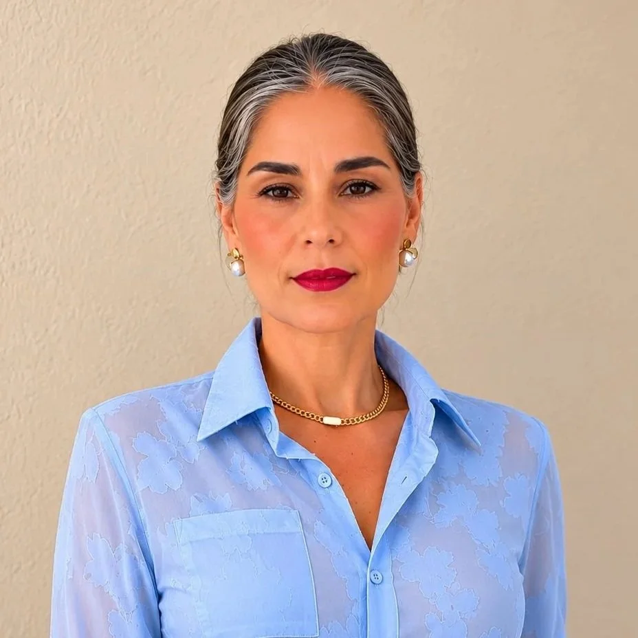A woman with gray hair, red lipstick, and pearl earrings wearing a light blue collared shirt standing against a beige wall.