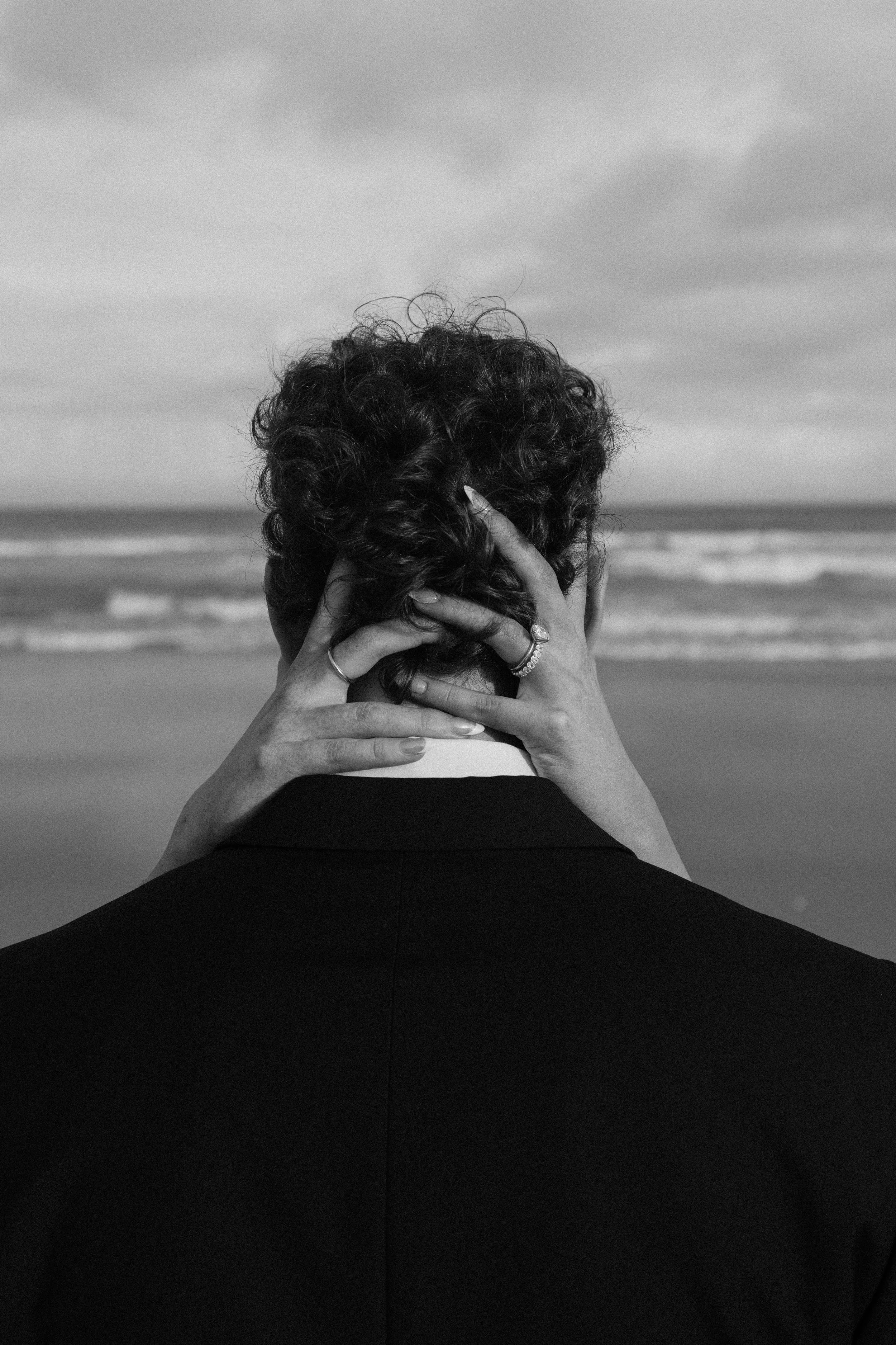 A person with curly hair holding their head in their hands at the beach, seen from behind, with ocean waves and cloudy sky in the background, in black and white.