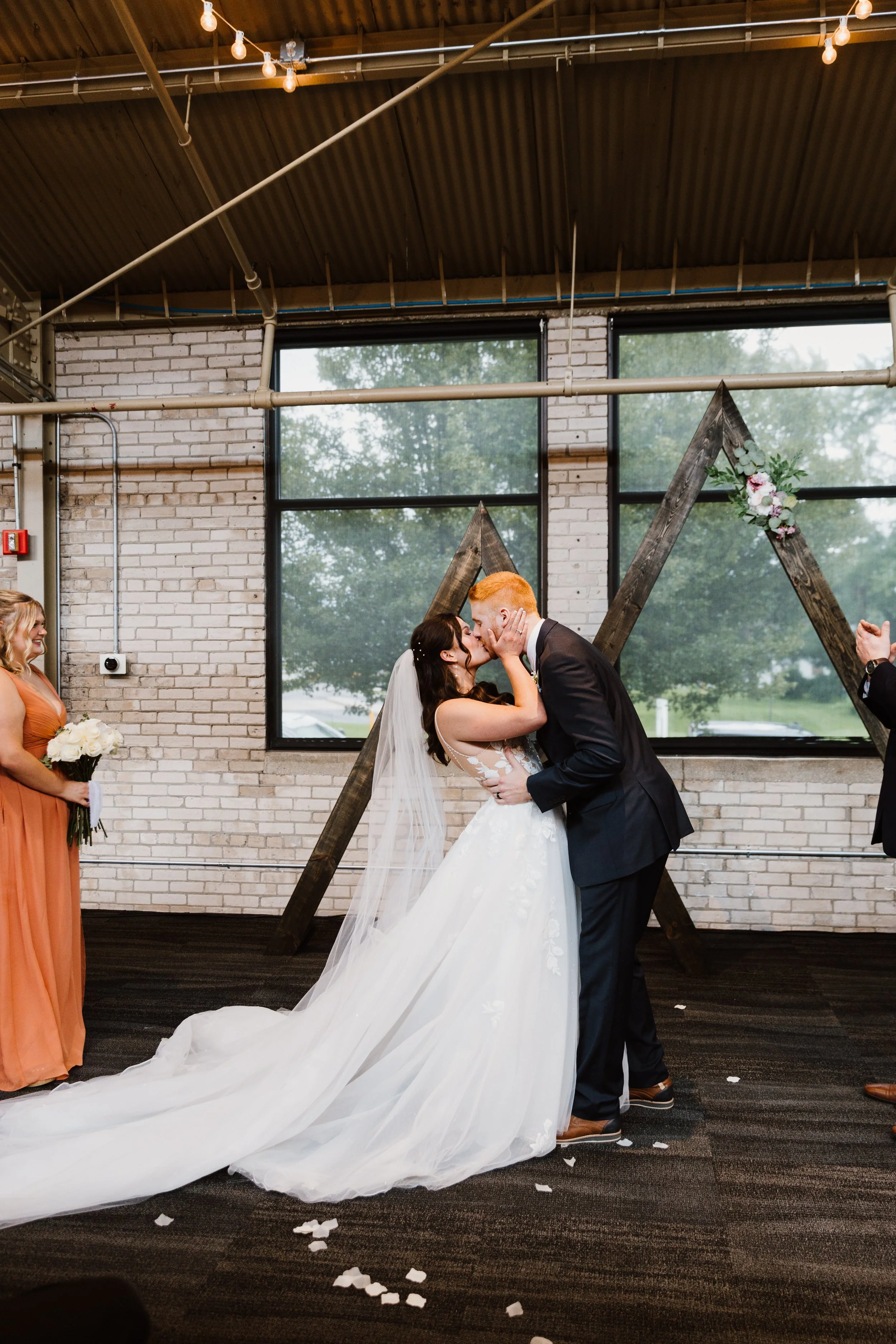 A bride and groom share a first kiss during their wedding ceremony indoors, with bridesmaid holding a bouquet on the side, and decorative wooden triangle with flowers in the background.