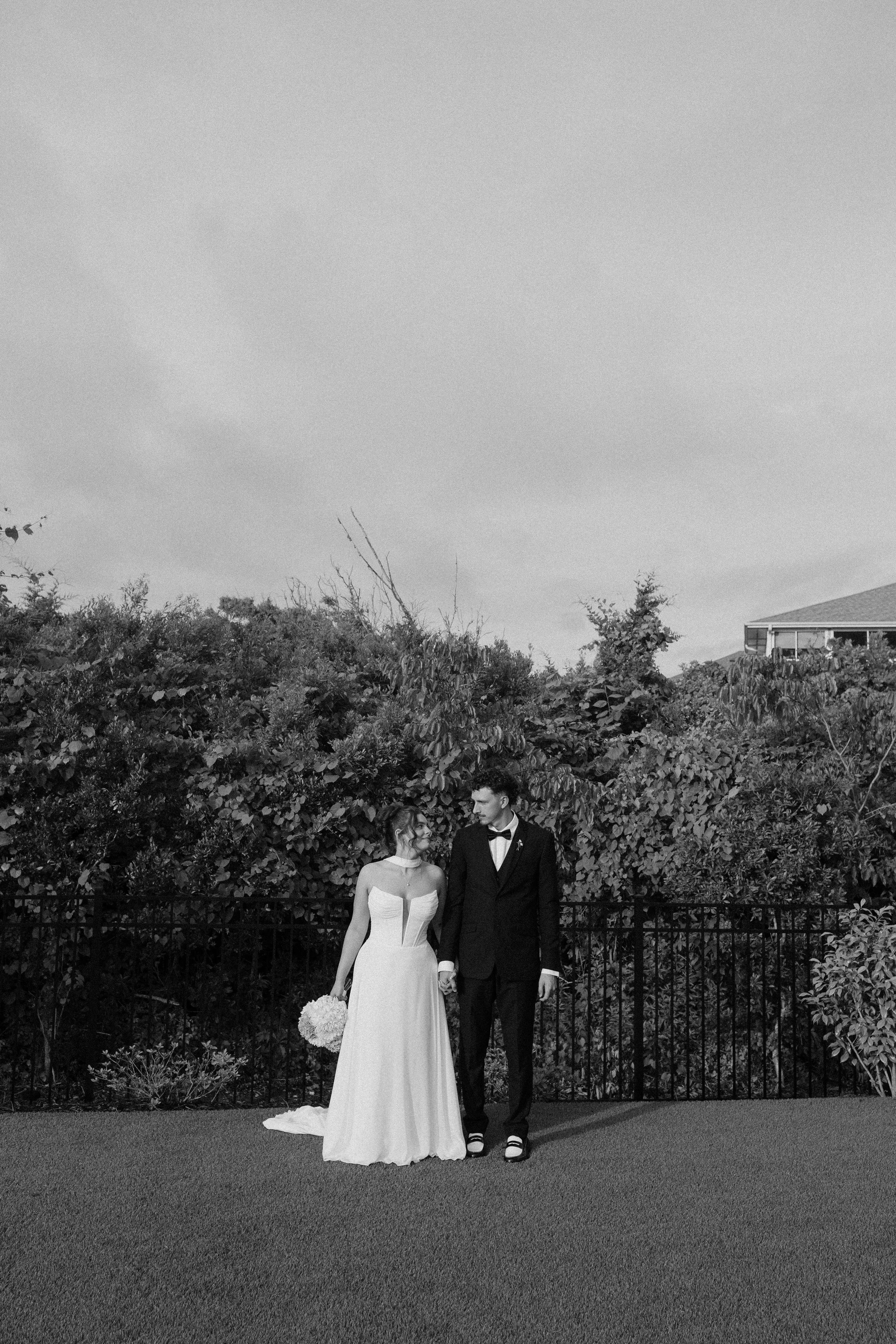 A bride and groom standing together outdoors during daytime, holding hands and looking at each other, with a lush green hedge and house in the background, black and white photograph.