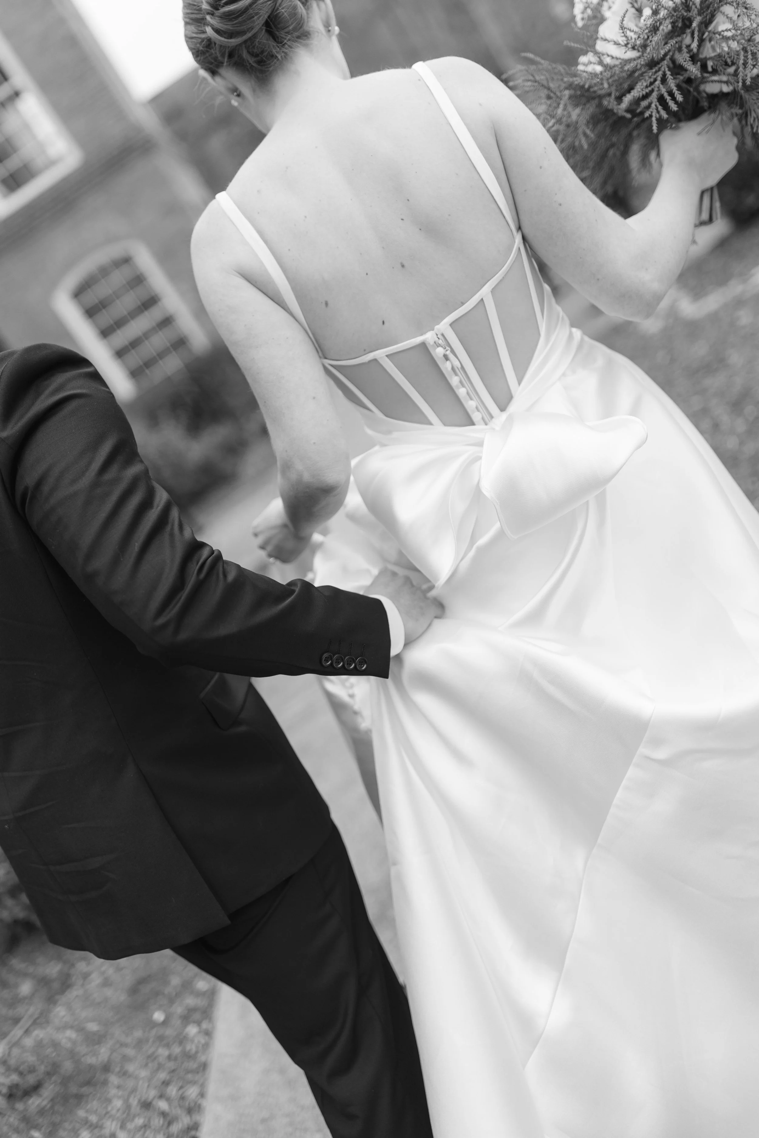 A bride and groom holding hands, with the bride wearing a wedding dress and the groom in a suit, outdoors in black and white.