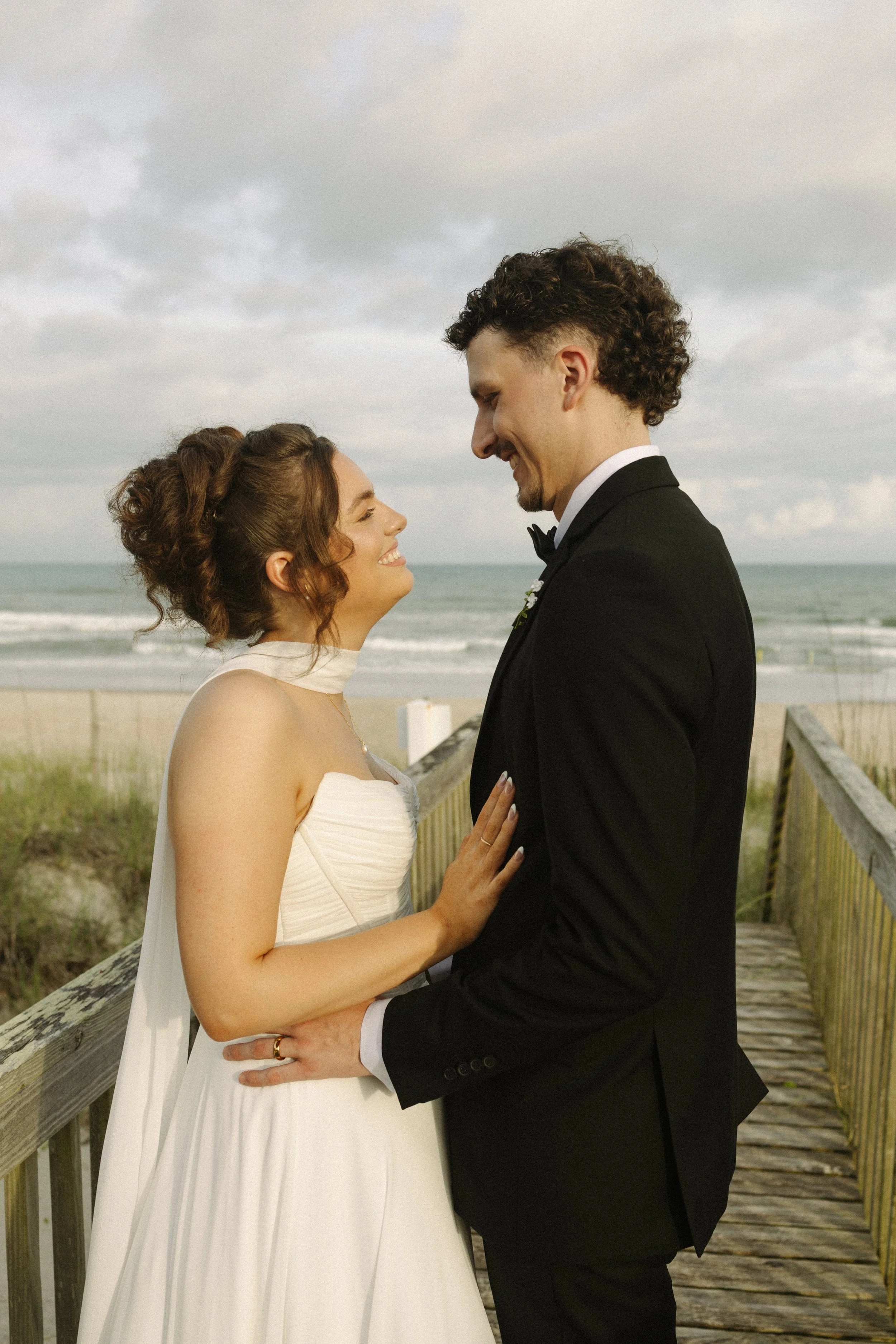 A couple dressed in wedding attire holding hands on a wooden pier at the beach, smiling and looking into each other's eyes with the ocean and cloudy sky in the background.