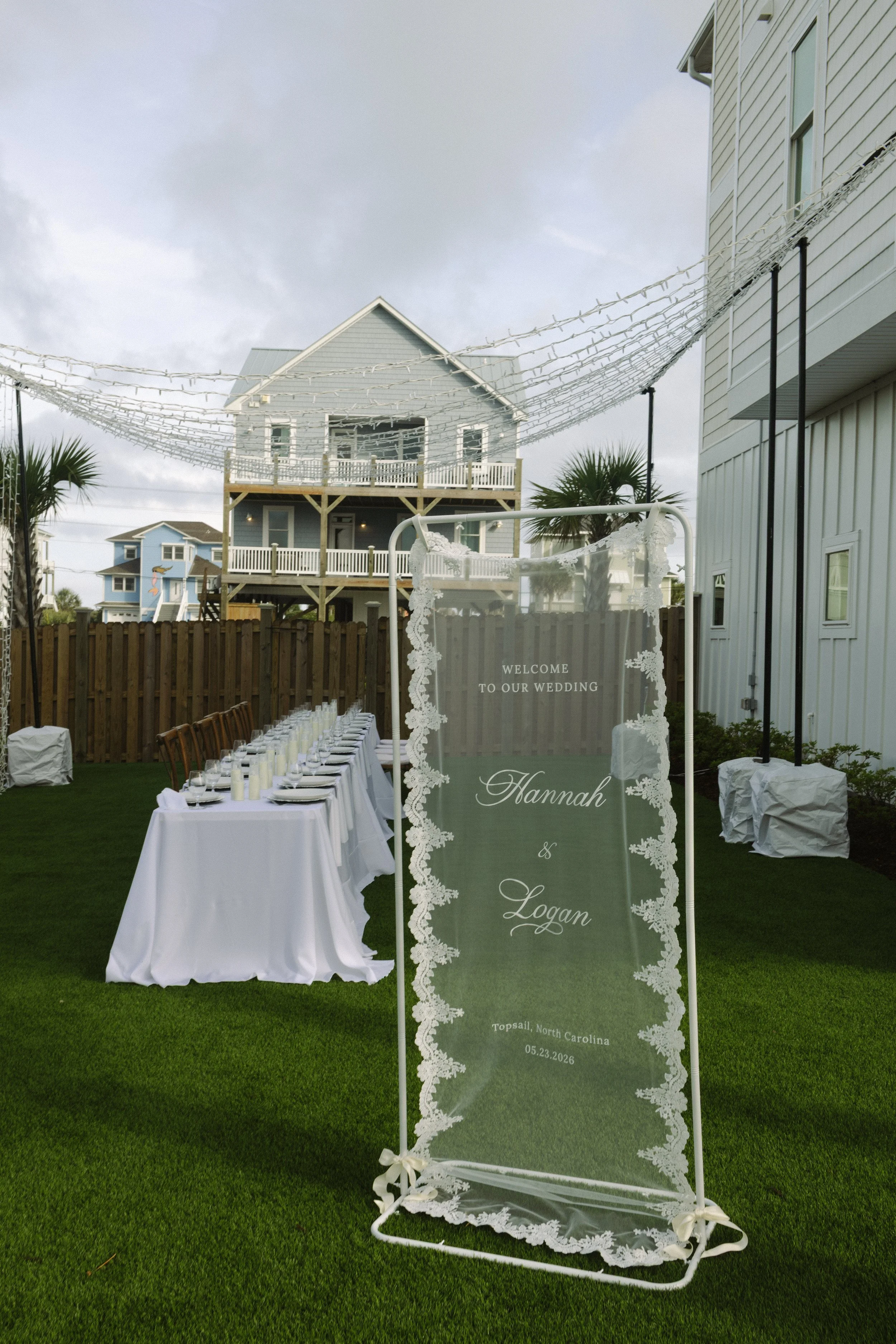 A wedding reception area set outdoors on green grass. There is a tall, clear sign with lace borders that says 'Welcome to our wedding, Hannah & Logan, Topsail, North Carolina, 05.23.2026'. In the background, there are wedding tables with white tablec