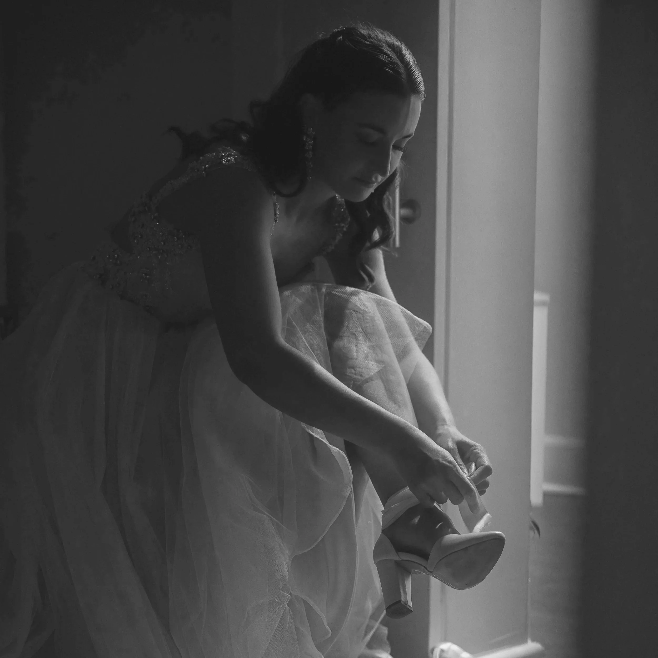 A black and white photo of a young woman sitting on the floor, looking down with a gentle smile, dressed in a sparkly top and a tulle skirt, in a softly lit indoor setting.