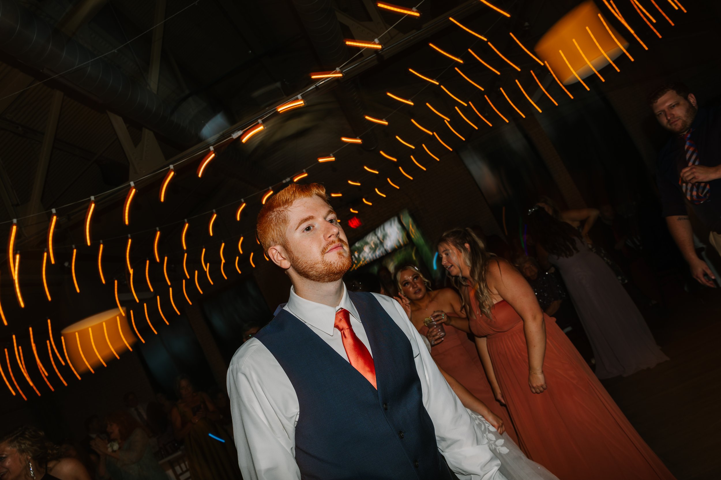 A young man with red hair and a beard, wearing a white shirt, navy vest, and red tie, standing in a dimly lit party with orange string lights overhead. Other guests are visible in the background, some talking and smiling.
