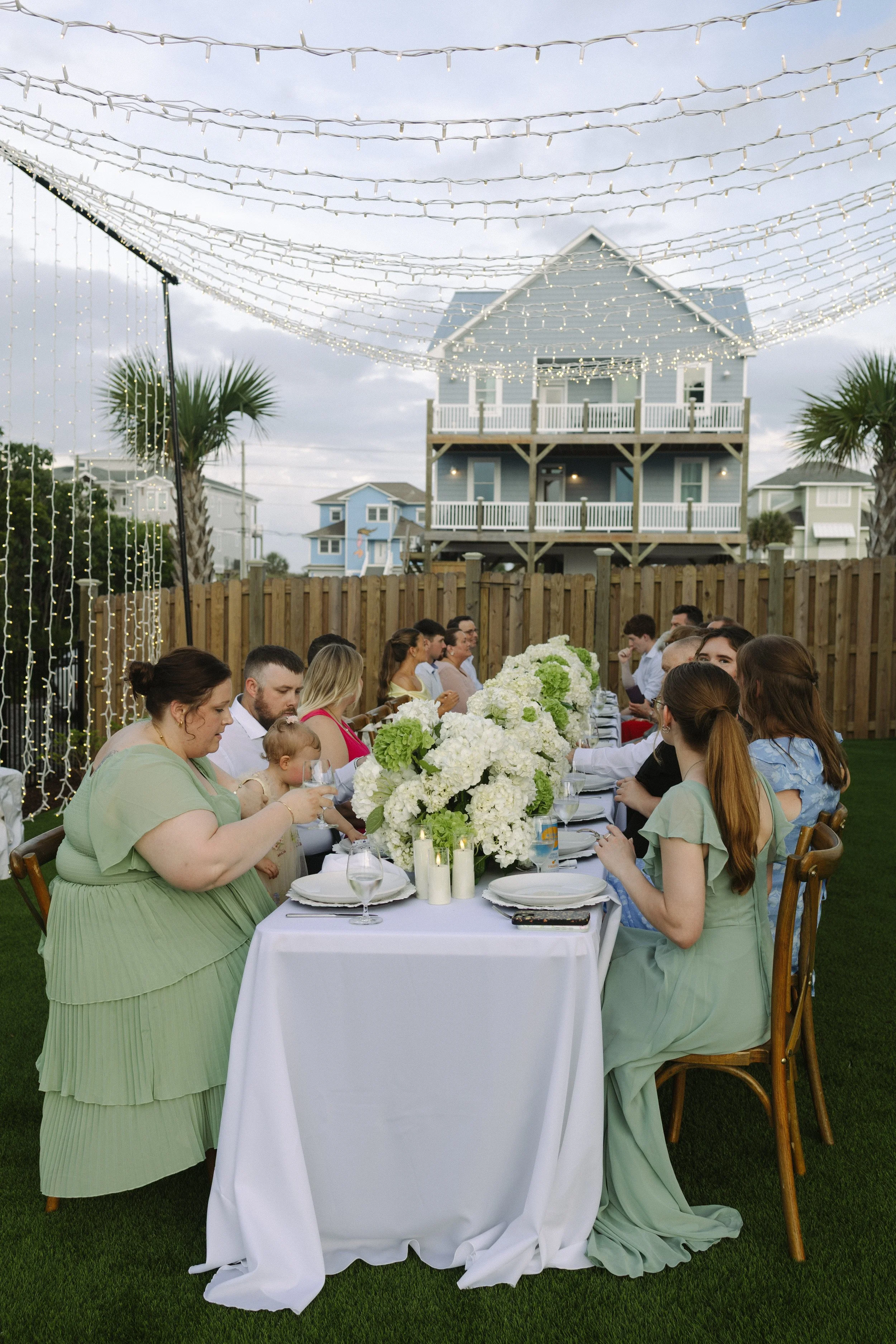 People gathered around a long outdoor dining table decorated with white flowers and candles, under string lights, in a backyard with palm trees and pastel-colored houses in the background.