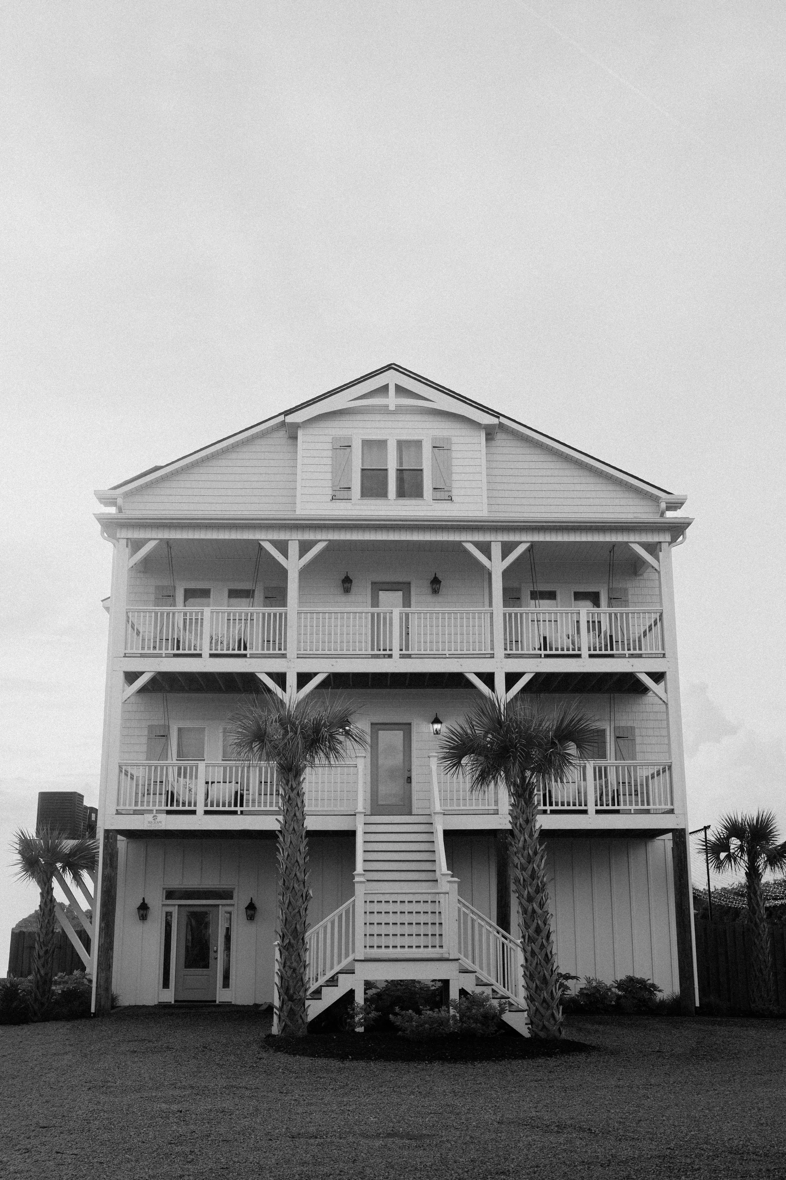 A three-story white house with a front porch, railing, and stairs, with three palm trees in front, and a gravel yard.