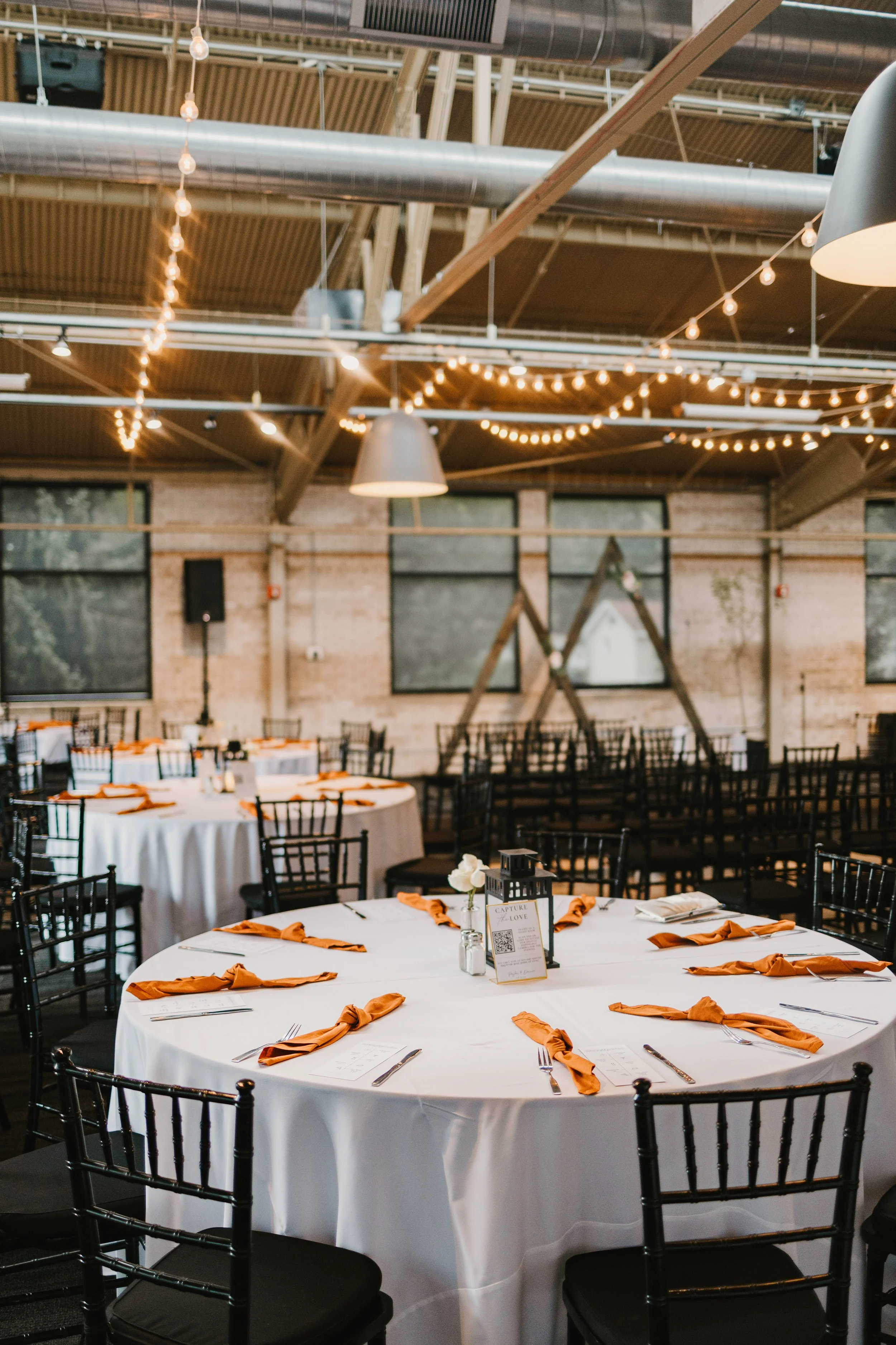 Decorated round banquet tables with white tablecloths, orange napkins tied in knots, and black chairs in a rustic event space with string lights and large windows.