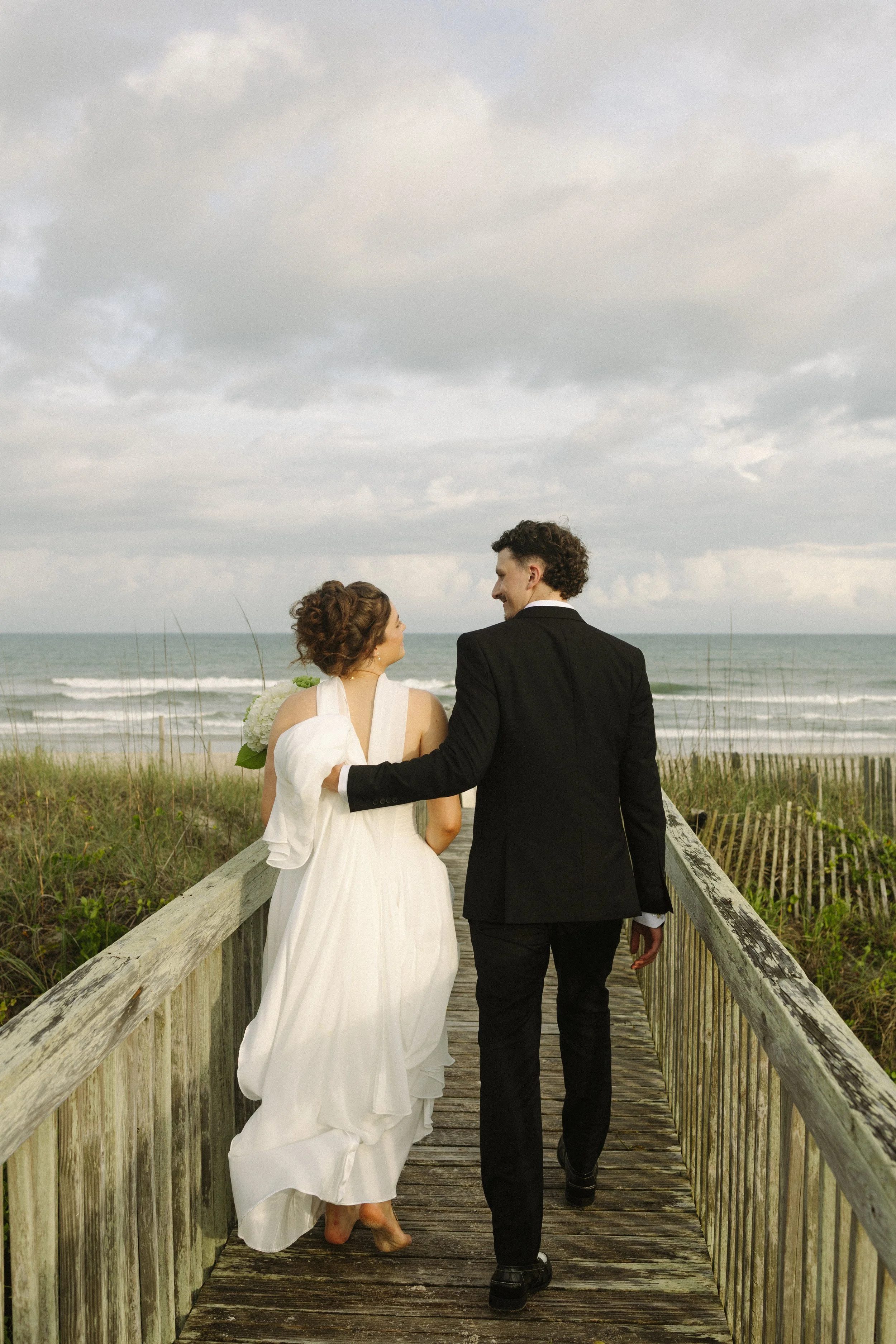 A bride and groom walking on a wooden boardwalk towards the beach, holding hands and smiling at each other, with ocean waves and cloudy sky in the background.