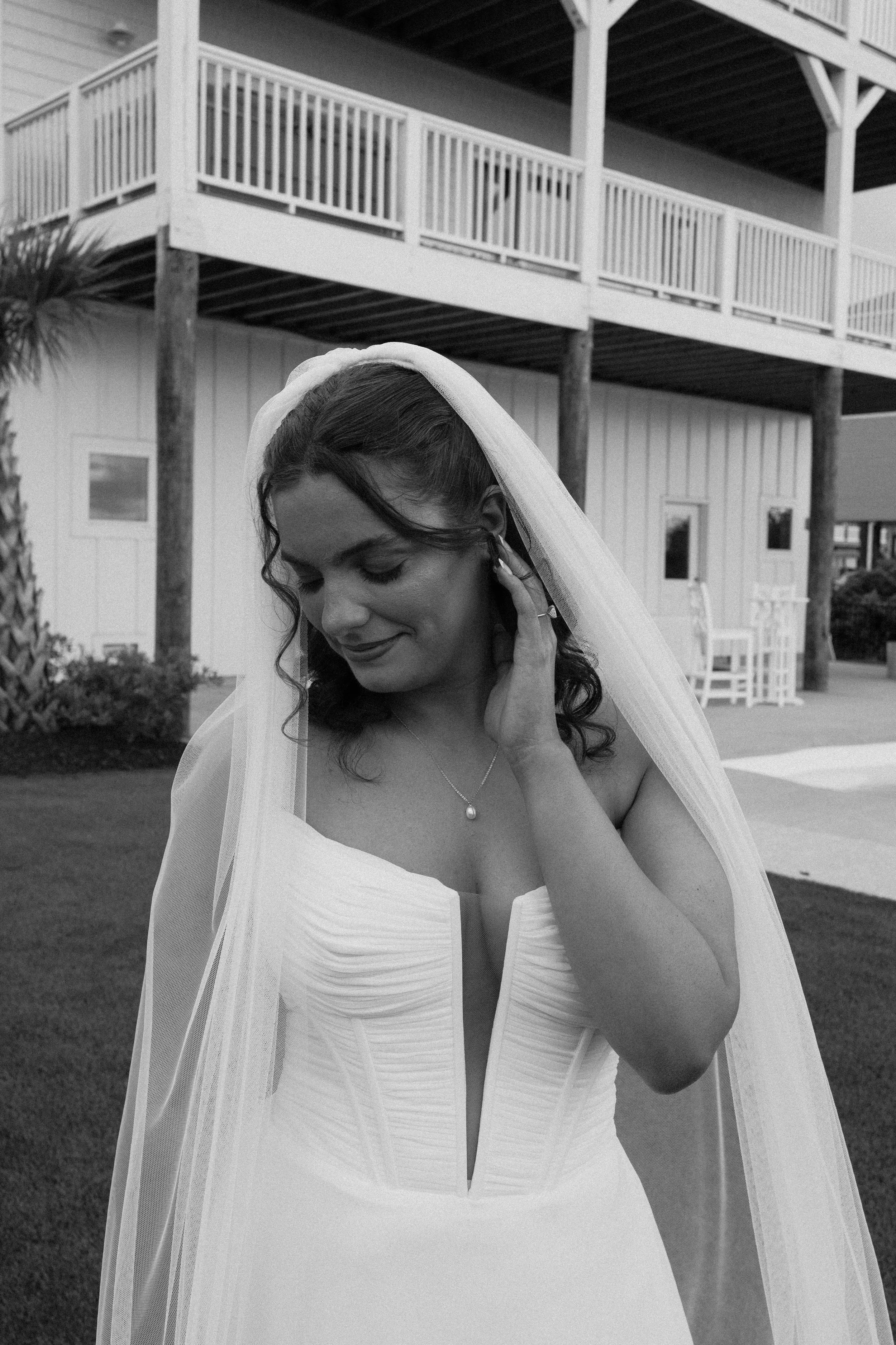 A bride with dark, curly hair wearing a strapless white wedding gown, a veil, and a pearl necklace, standing outdoors with a house and palm tree in the background.