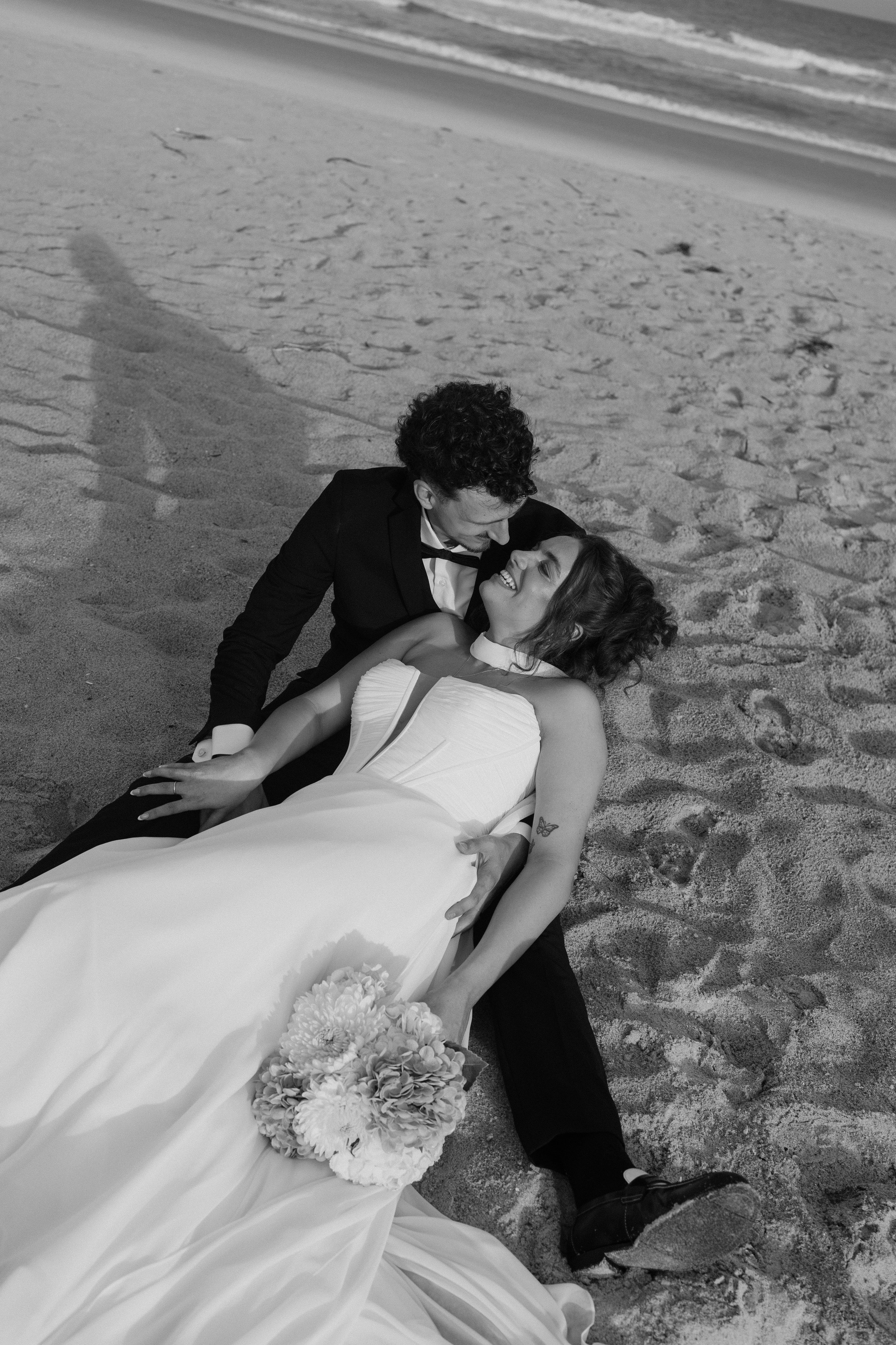A man and woman on the beach, dressed in wedding attire, with the man leaning over the woman who is lying on the sand holding a bouquet of flowers, smiling, in black and white.