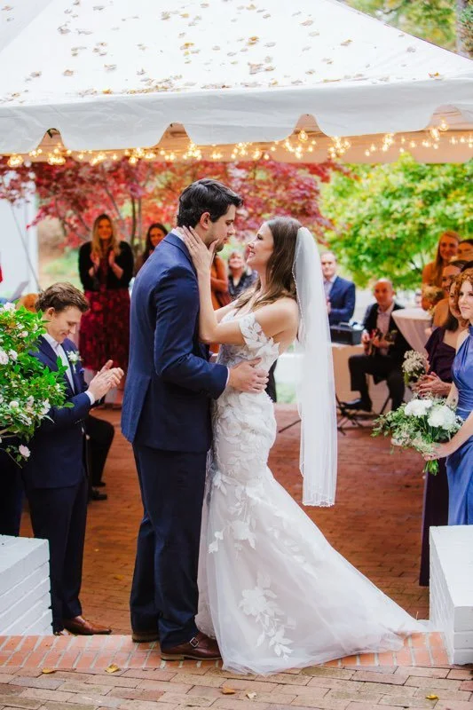 Bride and groom share a kiss during their wedding ceremony under a white canopy decorated with string lights, surrounded by seated guests and autumn trees in Raleigh, North Carolina.