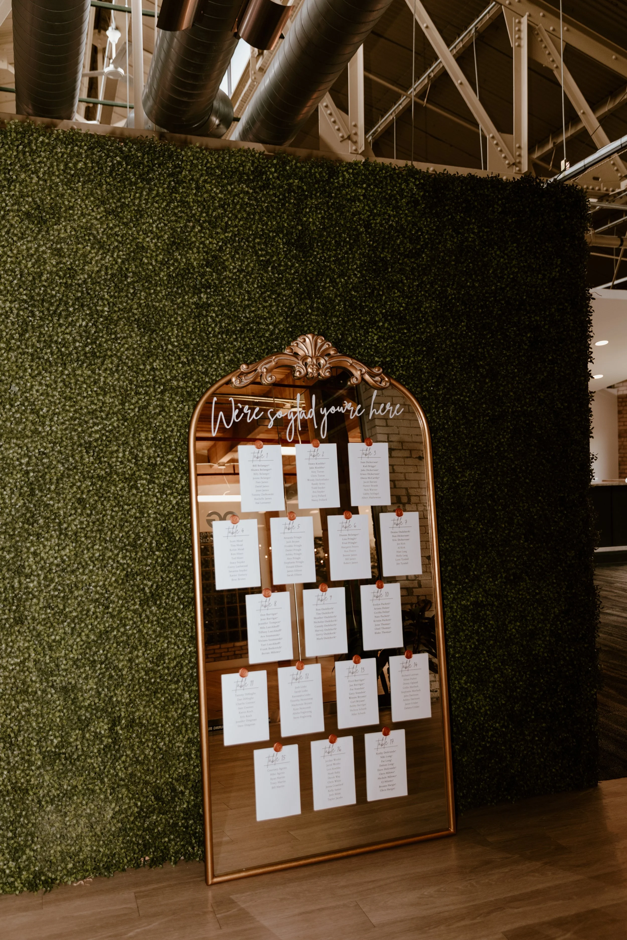 A decorative seating chart with a mirrored surface, floral border, and handwritten style text that says 'We're so glad you're here,' arranged in a grid on papers attached with clips, standing against a wall of green foliage.