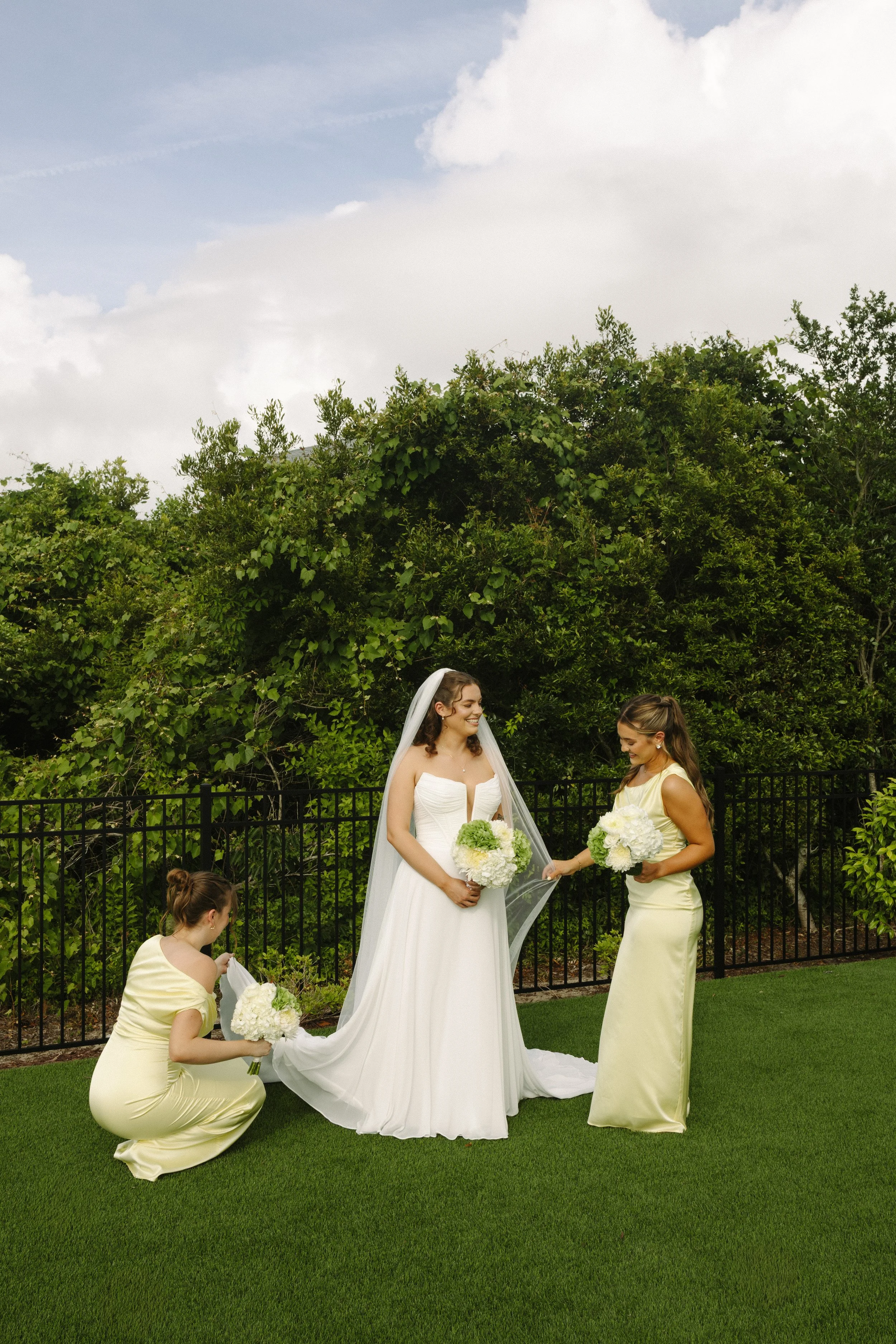 A bride in a white wedding dress and veil holding a bouquet of white and green flowers, standing on green grass with three women in yellow dresses. One woman is kneeling adjusting the wedding train, another is smiling and holding a bouquet, and the t