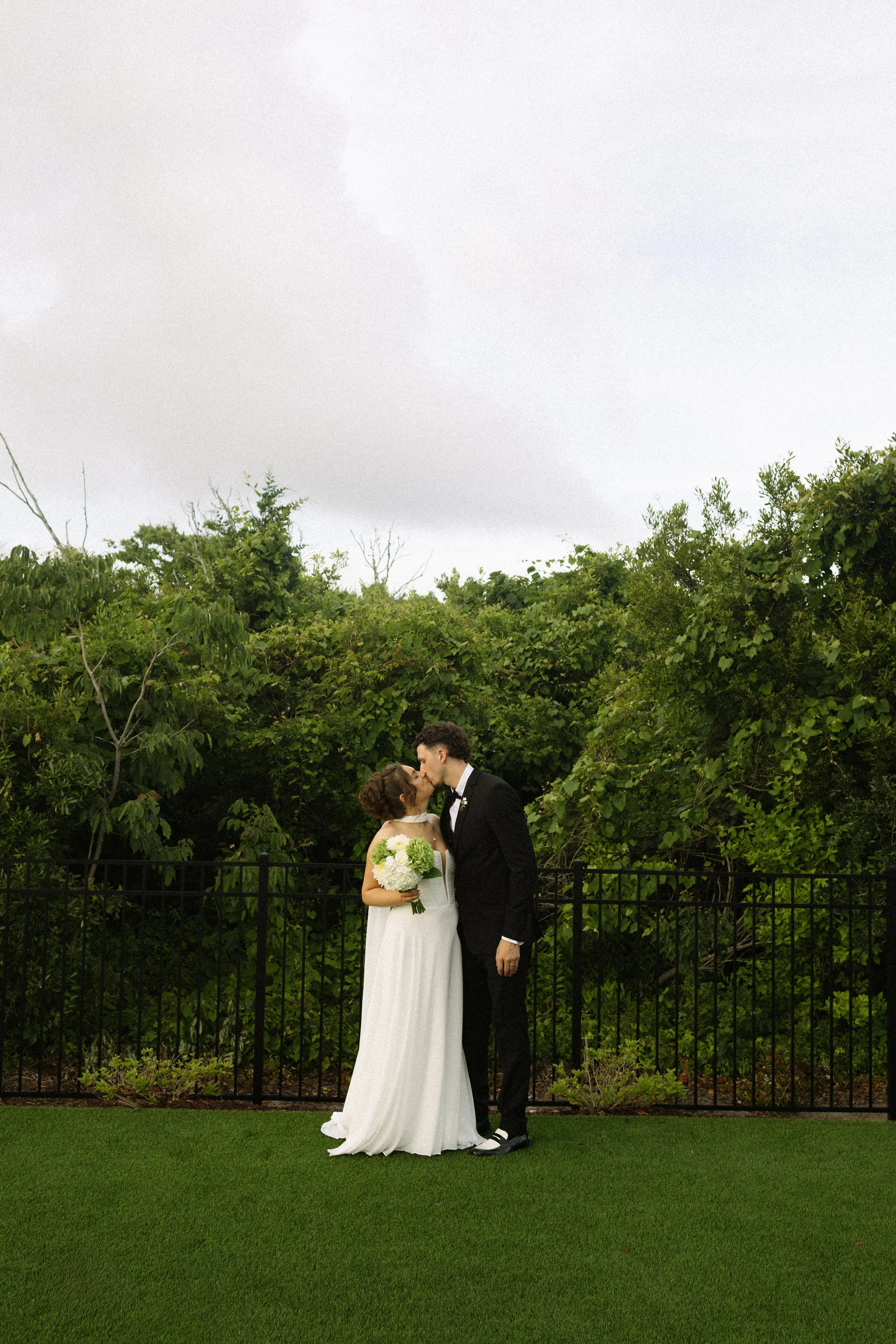 A bride and groom sharing a kiss outdoors during their wedding, with greenery and trees in the background and a black fence in front.