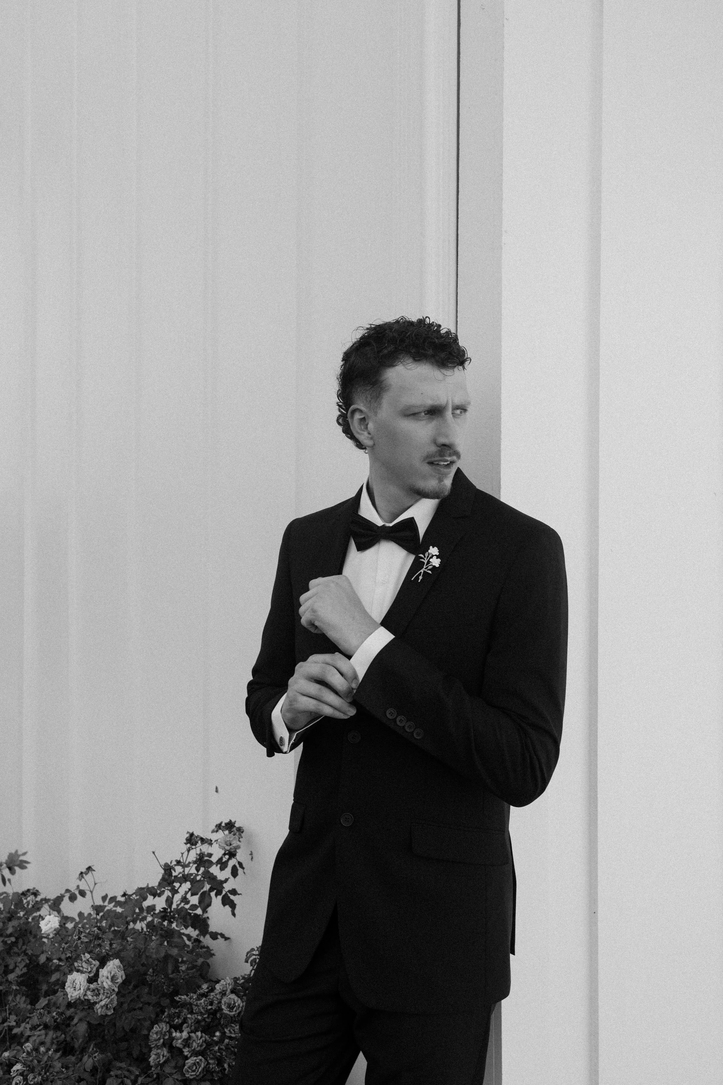 A man in a tuxedo with a bow tie standing indoors near a white wall and flowers, looking to his right with a serious expression.