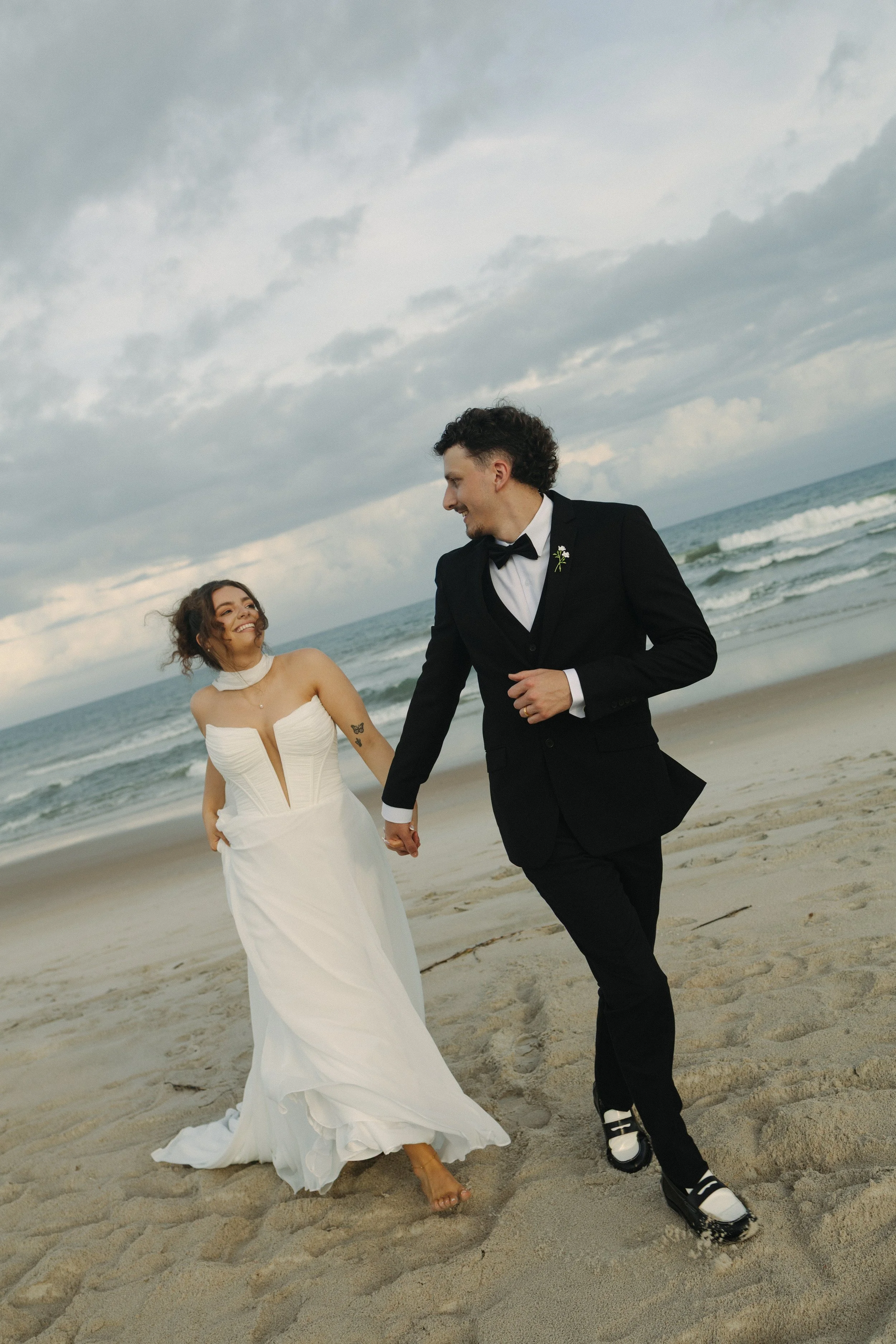 A couple in wedding attire walking hand in hand on a beach with waves and cloudy sky in the background.
