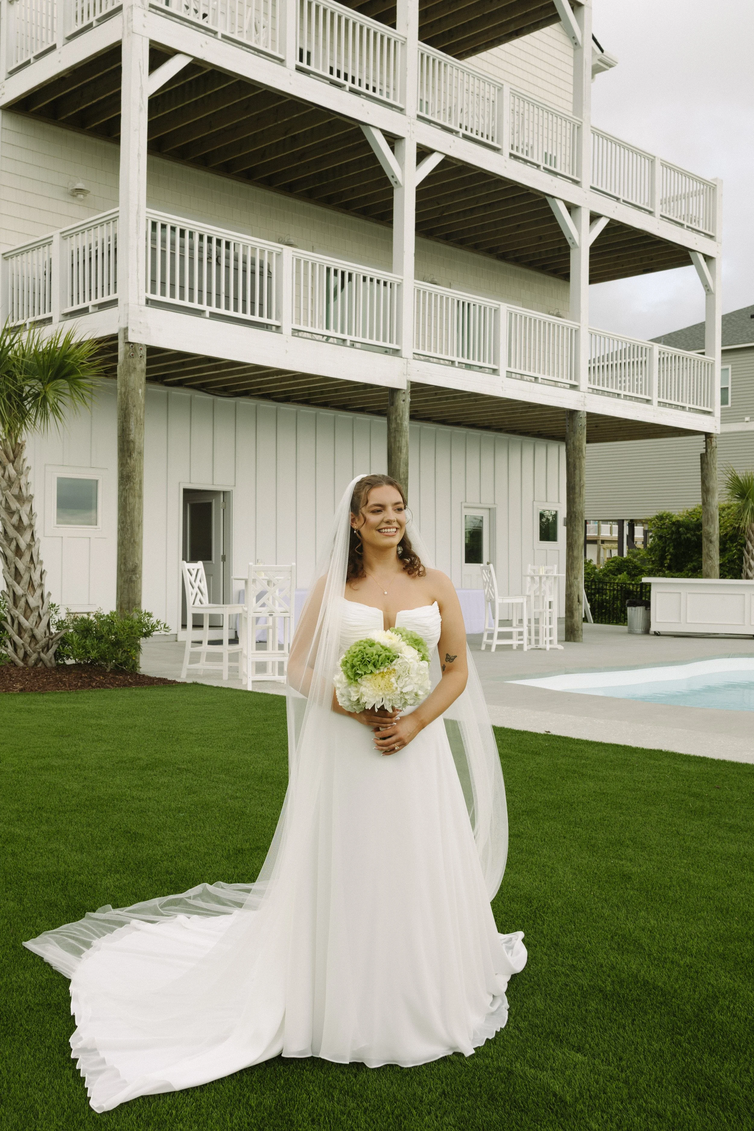 A bride in a strapless white wedding gown holding a bouquet of white and yellow flowers on a lush green lawn outside a two-story house with white siding and a large wooden deck.