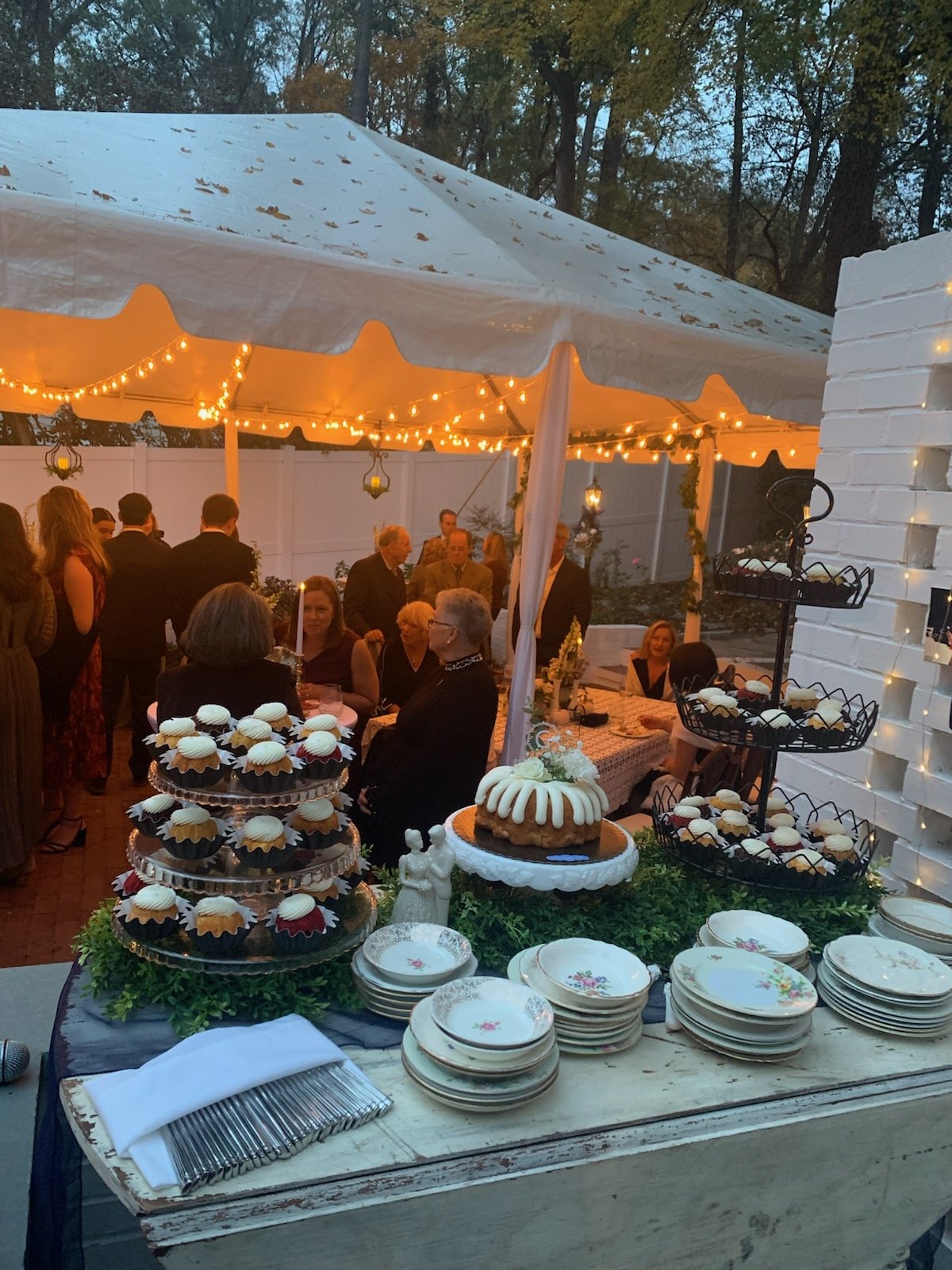 A dessert table at an outdoor gathering with cupcakes and a cake, set under a lit tent with guests mingling in the background in Raleigh, North Carolina..
