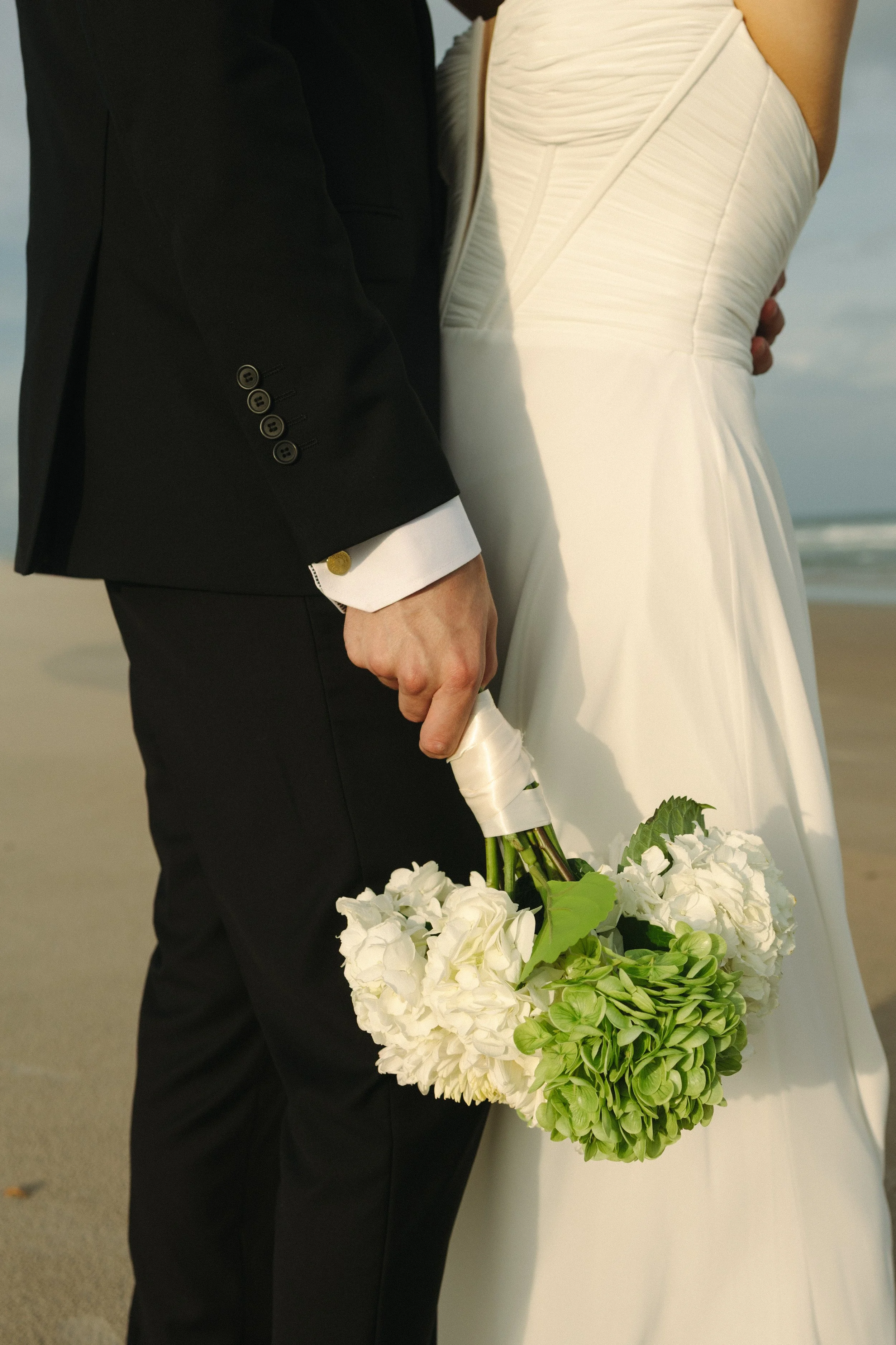A close-up of a bride and groom at the beach, with the bride holding a bouquet of white flowers and green leaves.