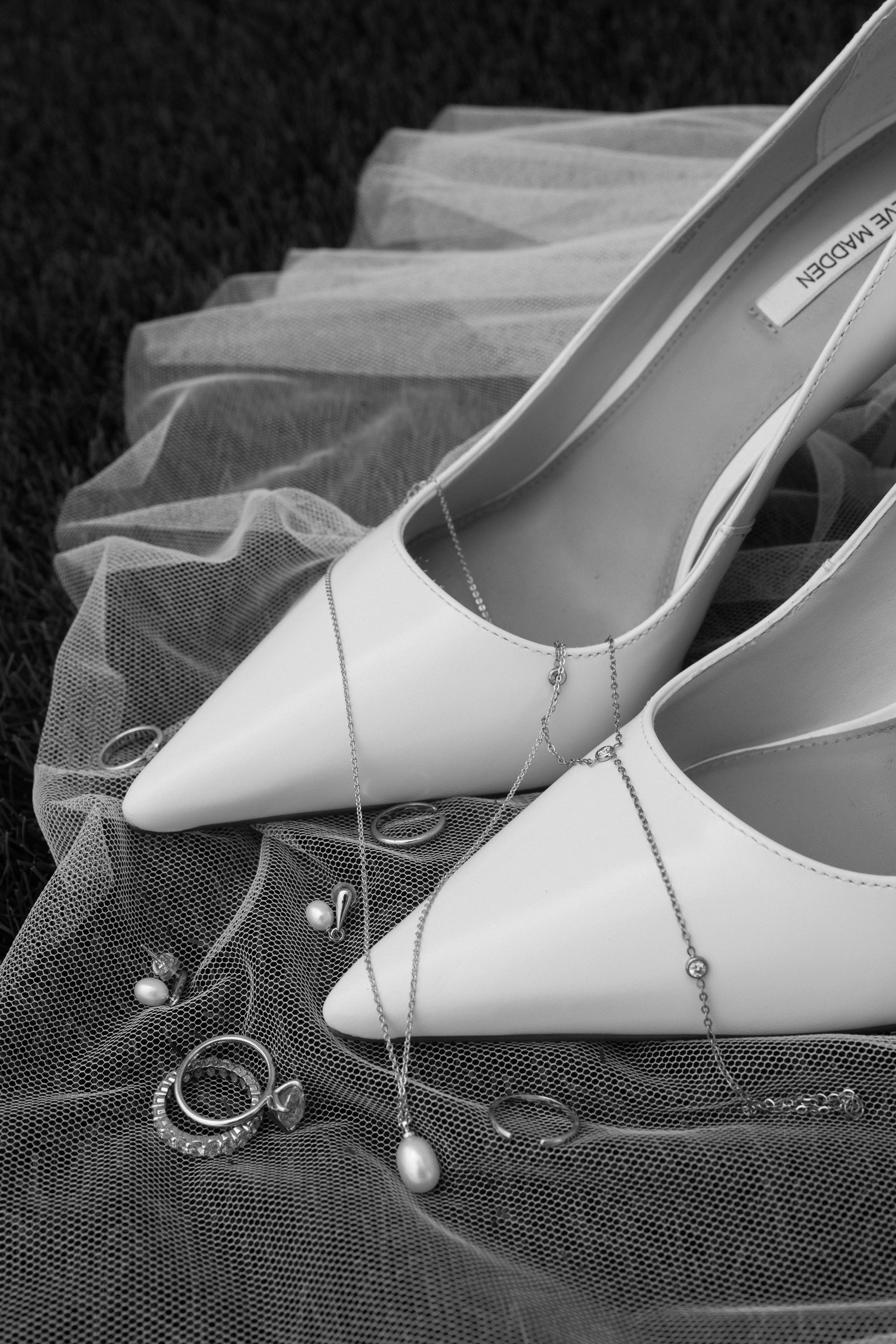 White high-heeled shoes and various jewelry pieces, including rings, earrings, and necklaces, arranged on a sheer fabric with a dark textured background.