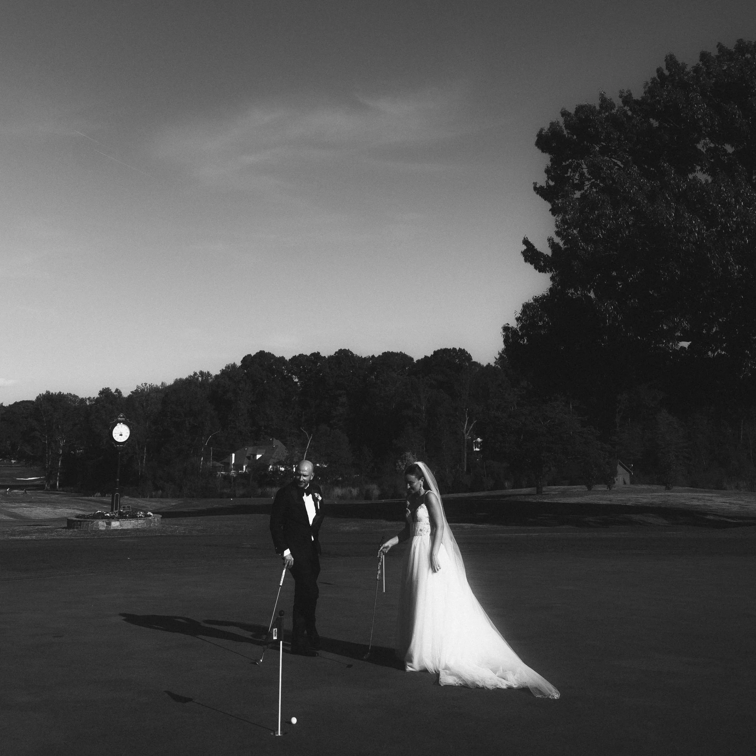 A bride and groom dressed in wedding attire playing mini golf outdoors at sunset, with trees and a clock tower visible in the background.