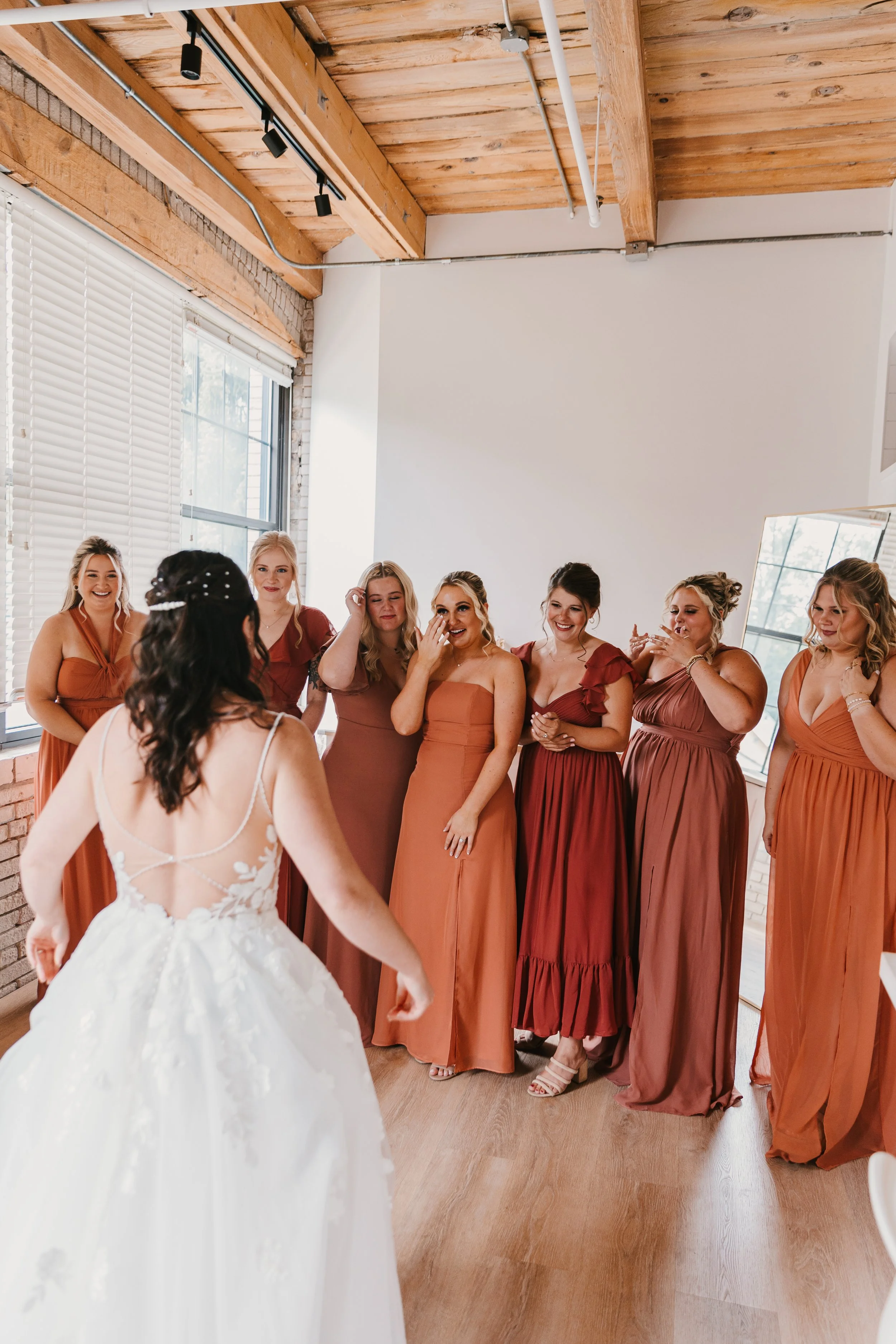 A bride in a white wedding dress is showing her back to seven bridesmaids in matching rust-colored dresses, who are reacting emotionally in a brightly lit room with wooden ceiling beams and large windows.