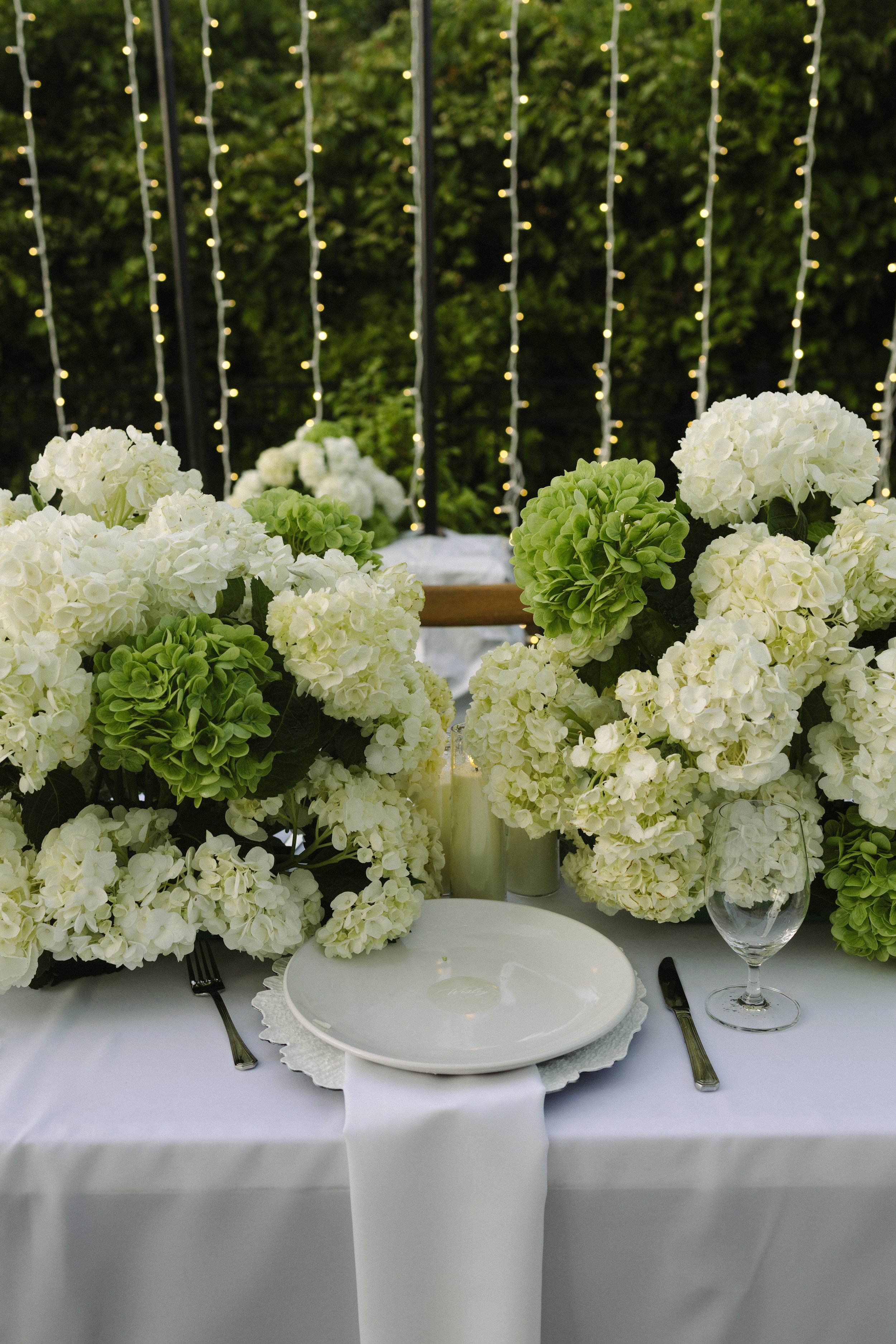 Elegant outdoor table setup with white hydrangea floral arrangements, white plates, a wine glass, silverware, and string lights in the background, suggesting a special occasion or celebration.