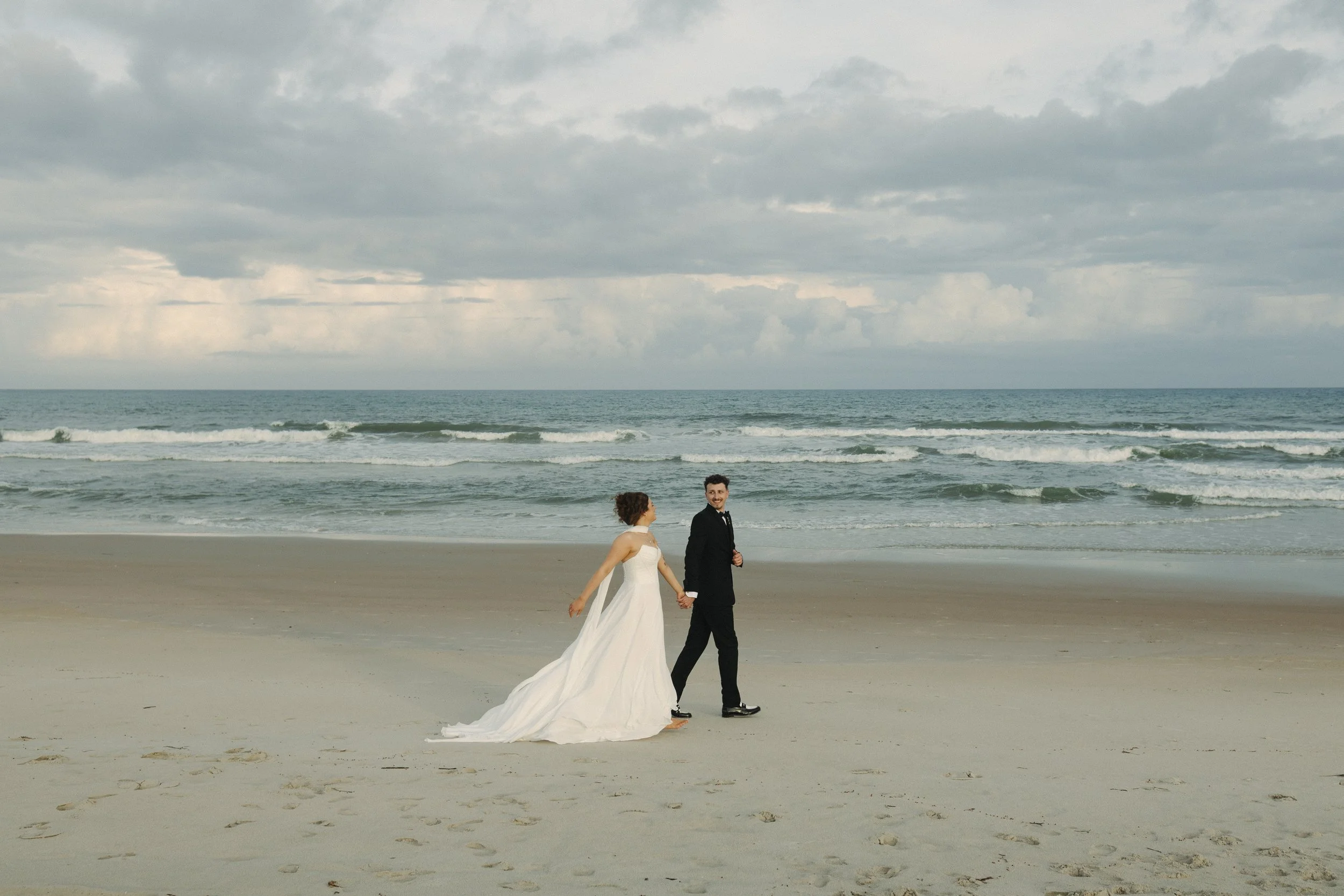 A bride and groom holding hands on a beach with the ocean and cloudy sky in the background.