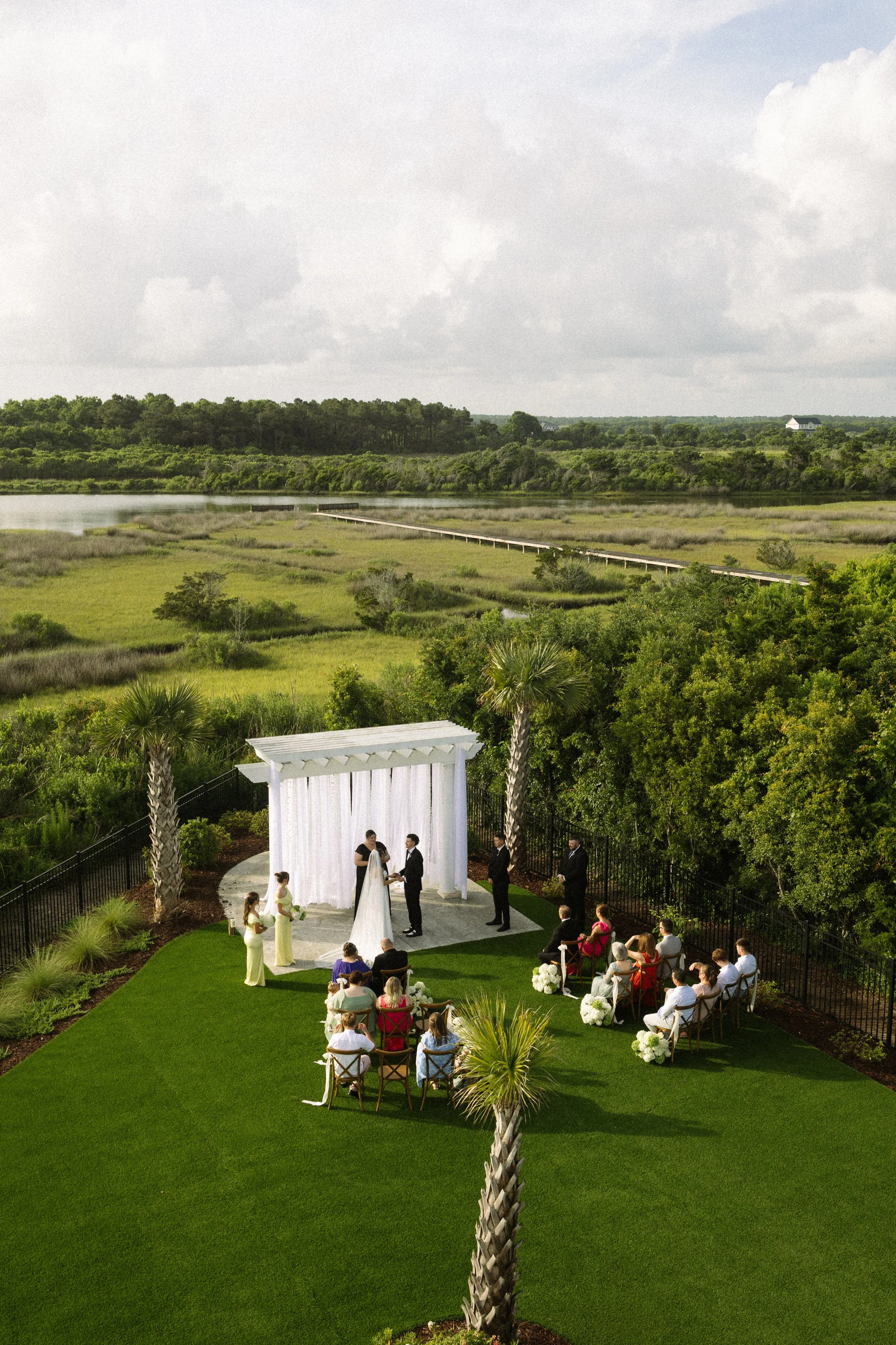 An outdoor wedding ceremony with a couple exchanging vows under a white arch, surrounded by family and friends, with lush greenery and a scenic landscape in the background.