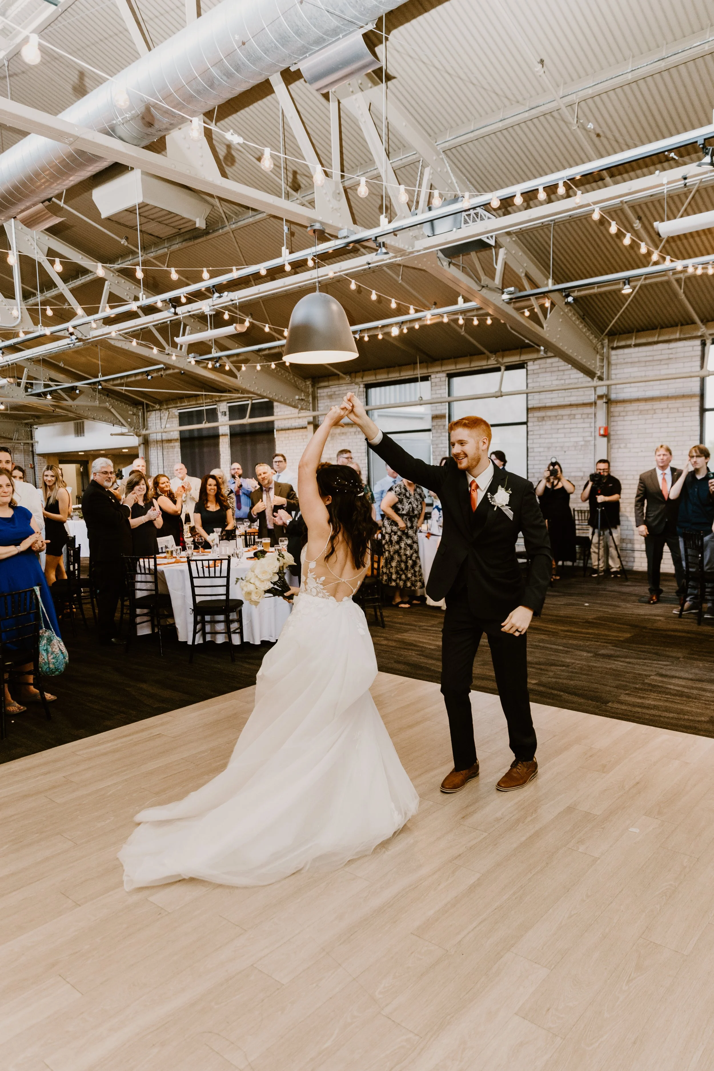 Bride and groom dancing at wedding reception with guests watching and taking photos in an industrial-style venue with string lights and brick walls.