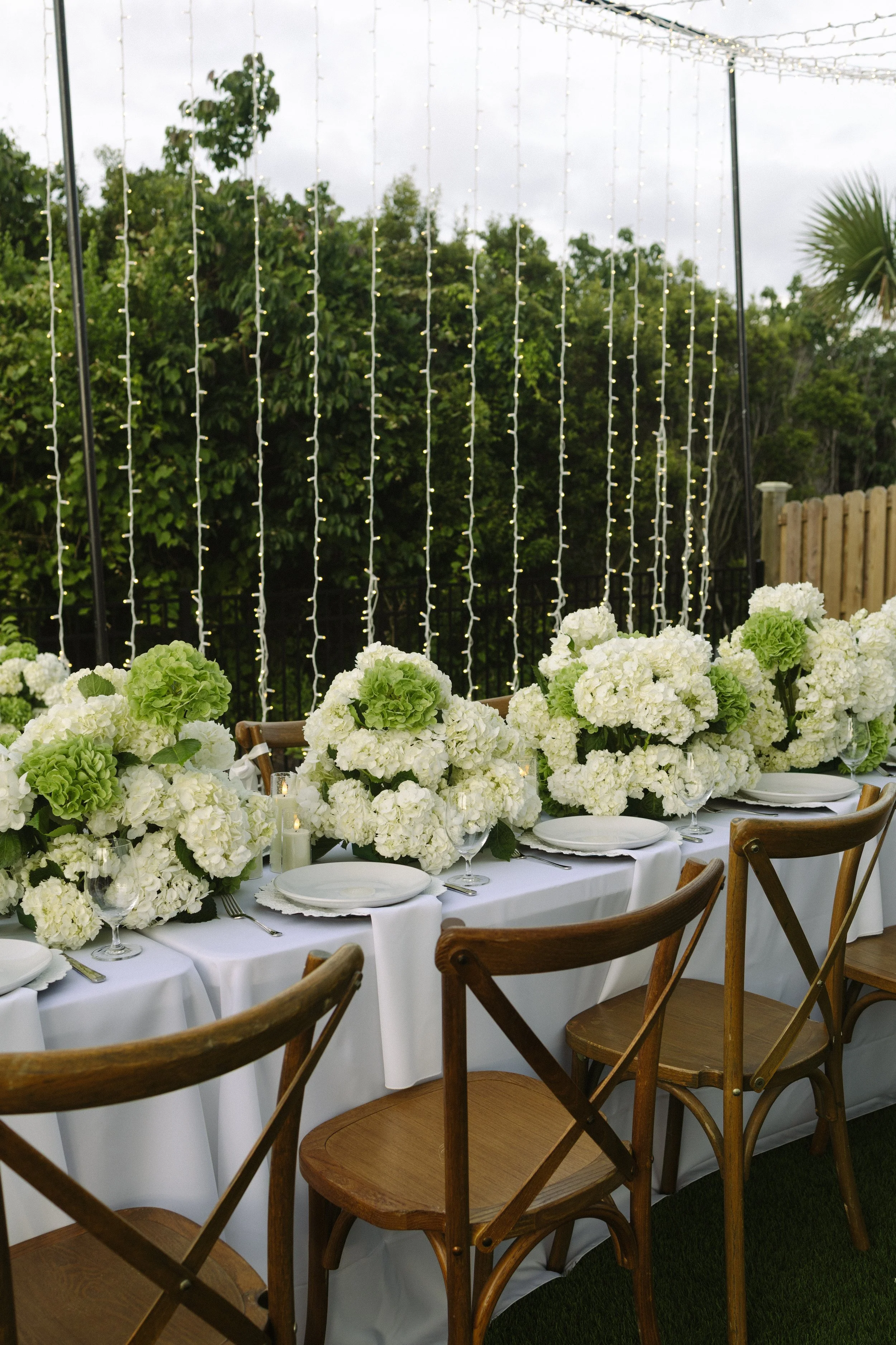 An outdoor dining table decorated with white hydrangea flowers and greenery, set with white plates and glassware, under a canopy of string lights with a forest in the background.