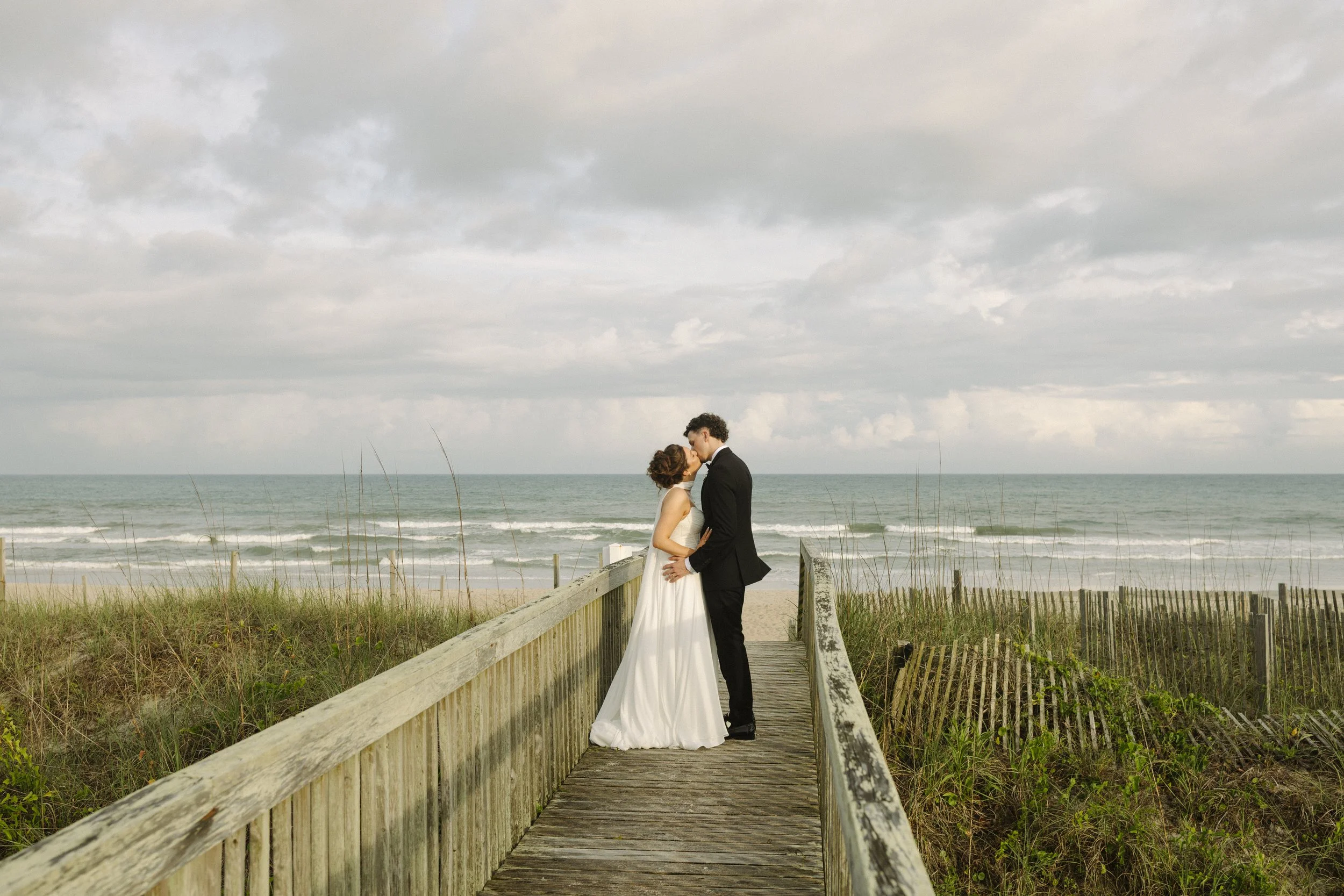 A couple in wedding attire embrace on a wooden boardwalk on the beach, with the ocean and cloudy sky in the background.