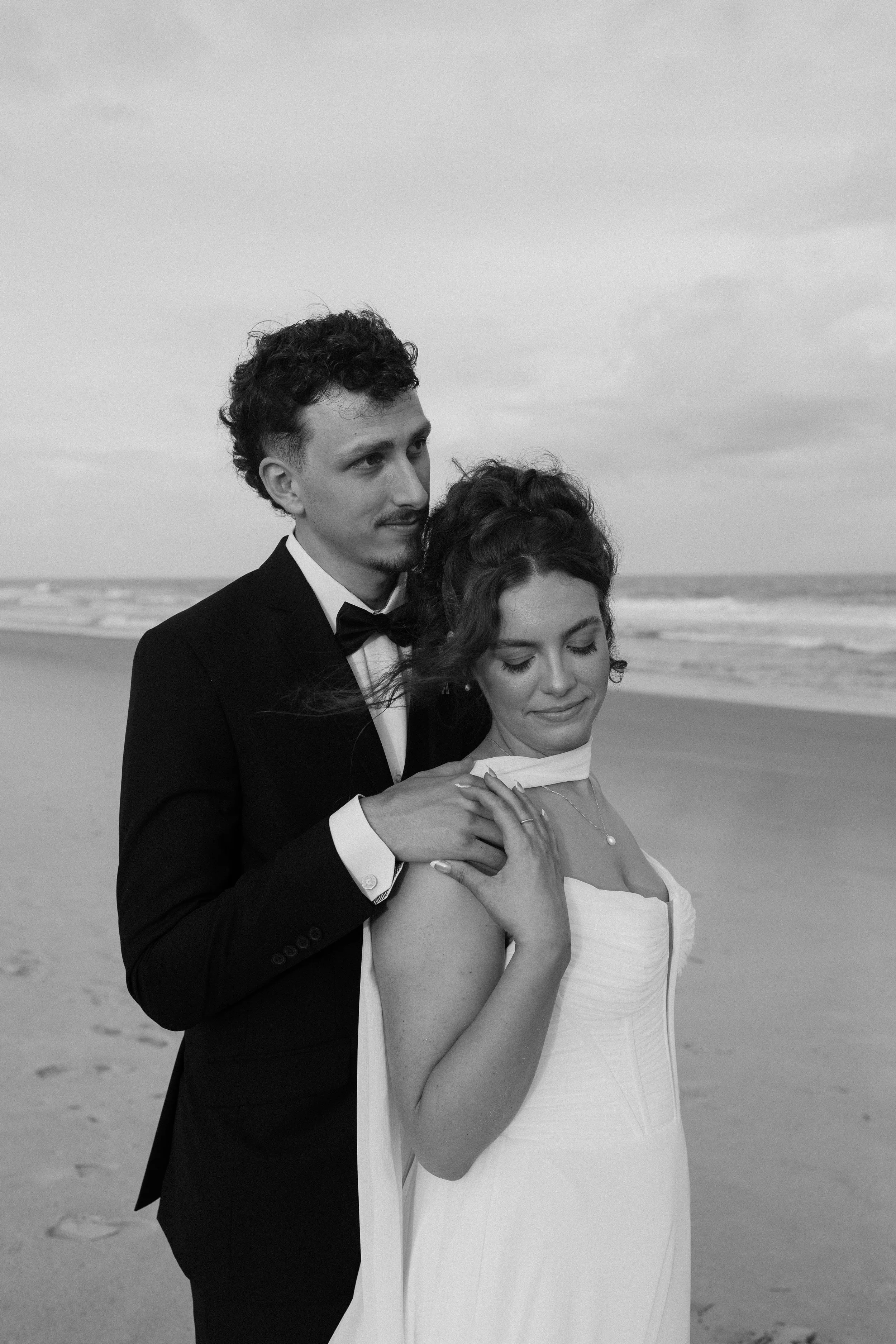 A man in a tuxedo with a bow tie and a woman in a wedding dress on the beach, with the ocean and cloudy sky in the background.
