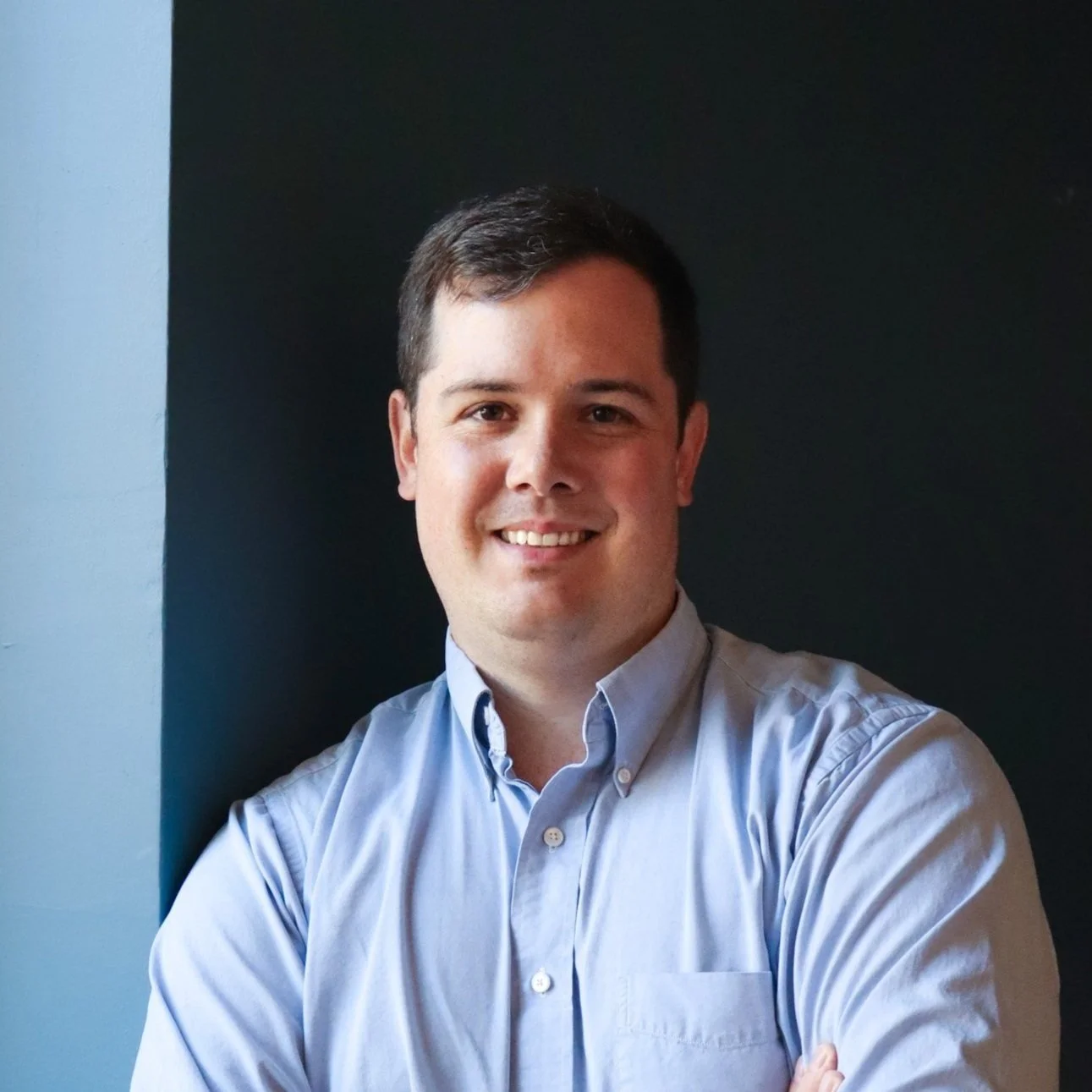 Somdal Associates architecture interior design, louisiana, shreveport, baton rouge, Portrait of a young man with dark hair, smiling, wearing a light blue button-up shirt, standing against a dark background.